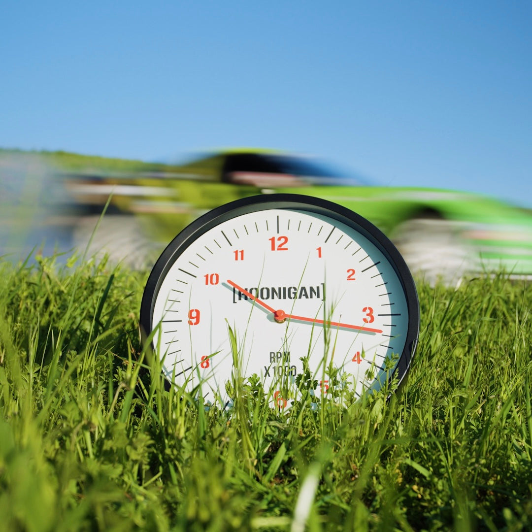 Hoonigan RPM Glow Clock in grass with a blurred race car in the background, showcasing automotive lifestyle and speed.