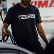 A person wearing a black Hoonigan AUTOBAHN short sleeve t-shirt refuels a car at a gas station.