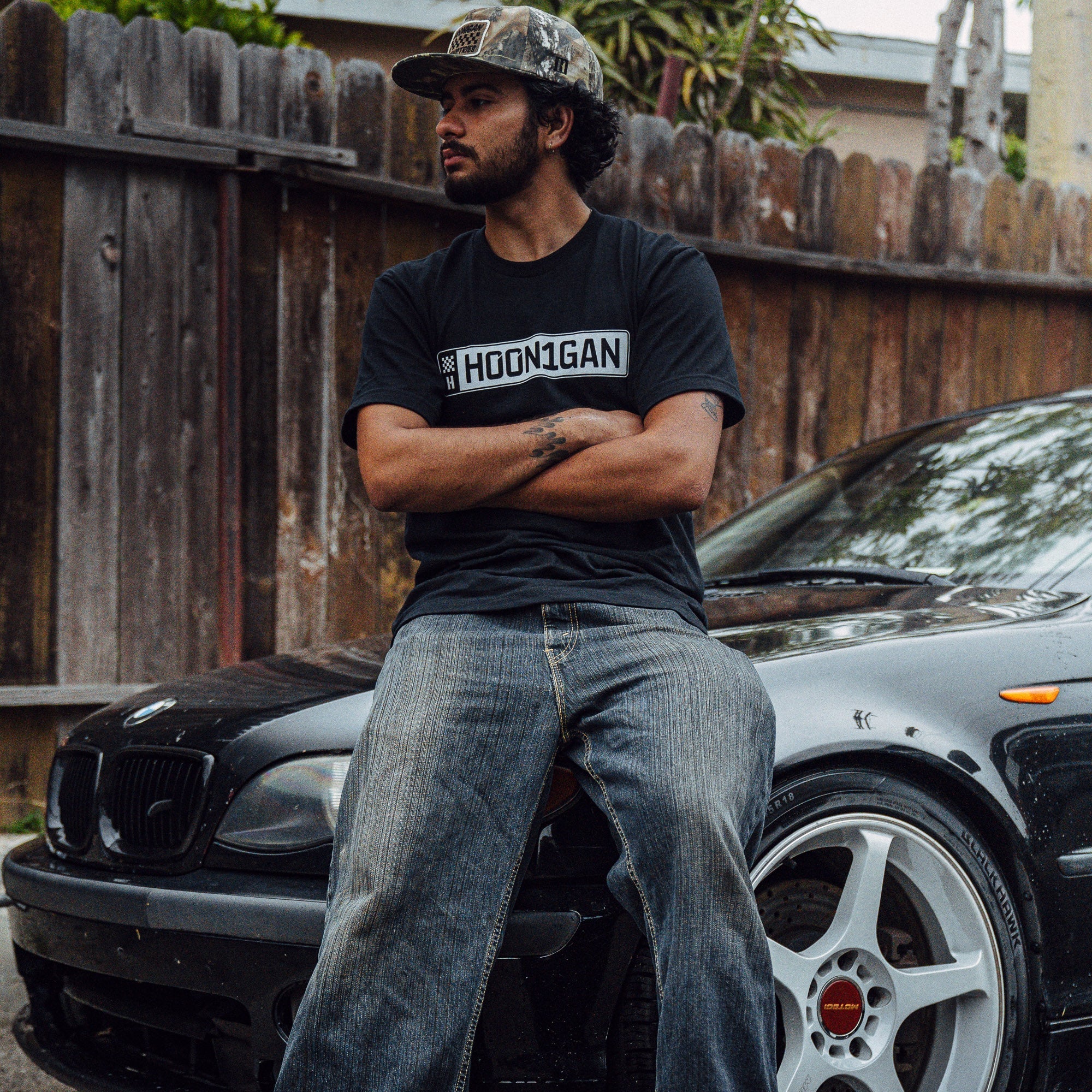 A person wearing a Hoonigan AUTOBAHN Short Sleeve T-Shirt sits on a car, showcasing automotive lifestyle and culture.