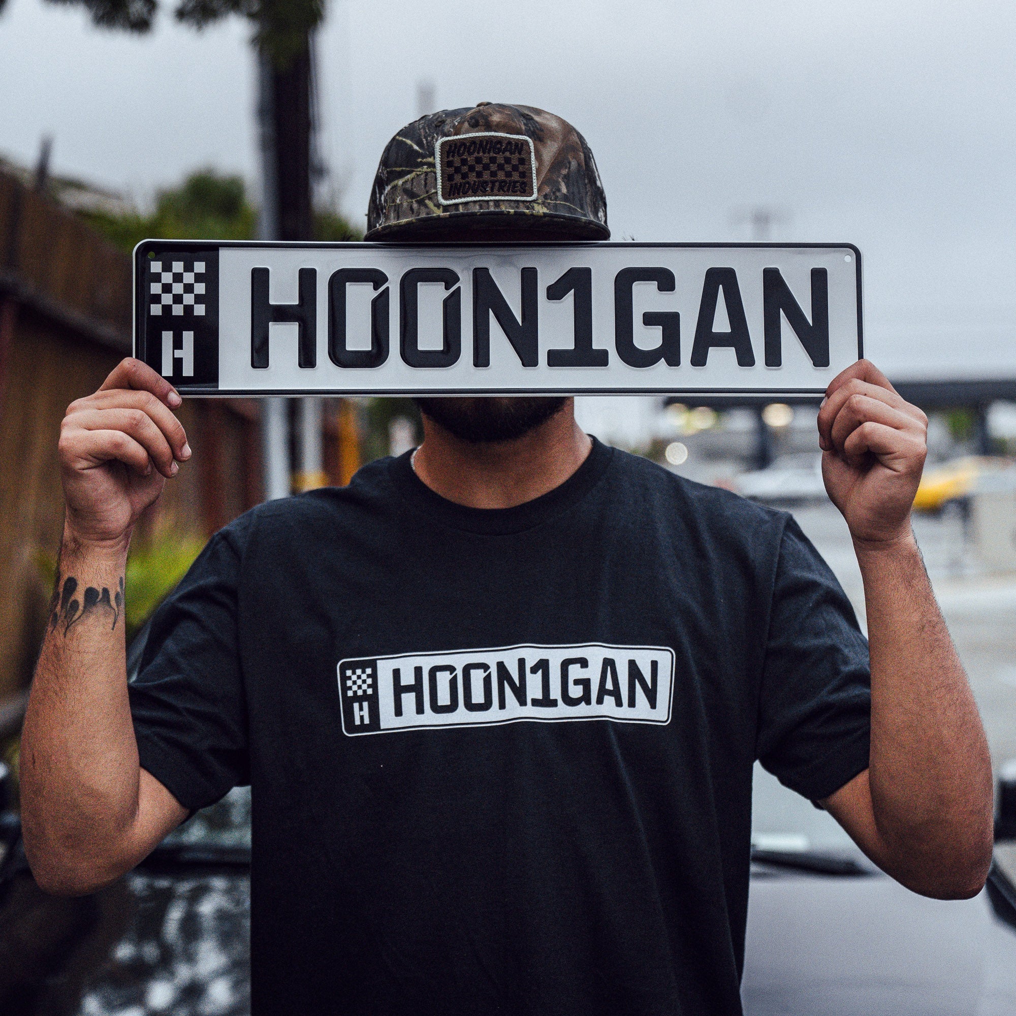 Man wearing a black Hoonigan AUTOBAHN short sleeve t-shirt, holding a license plate with "HOON1GAN" in front of his face.