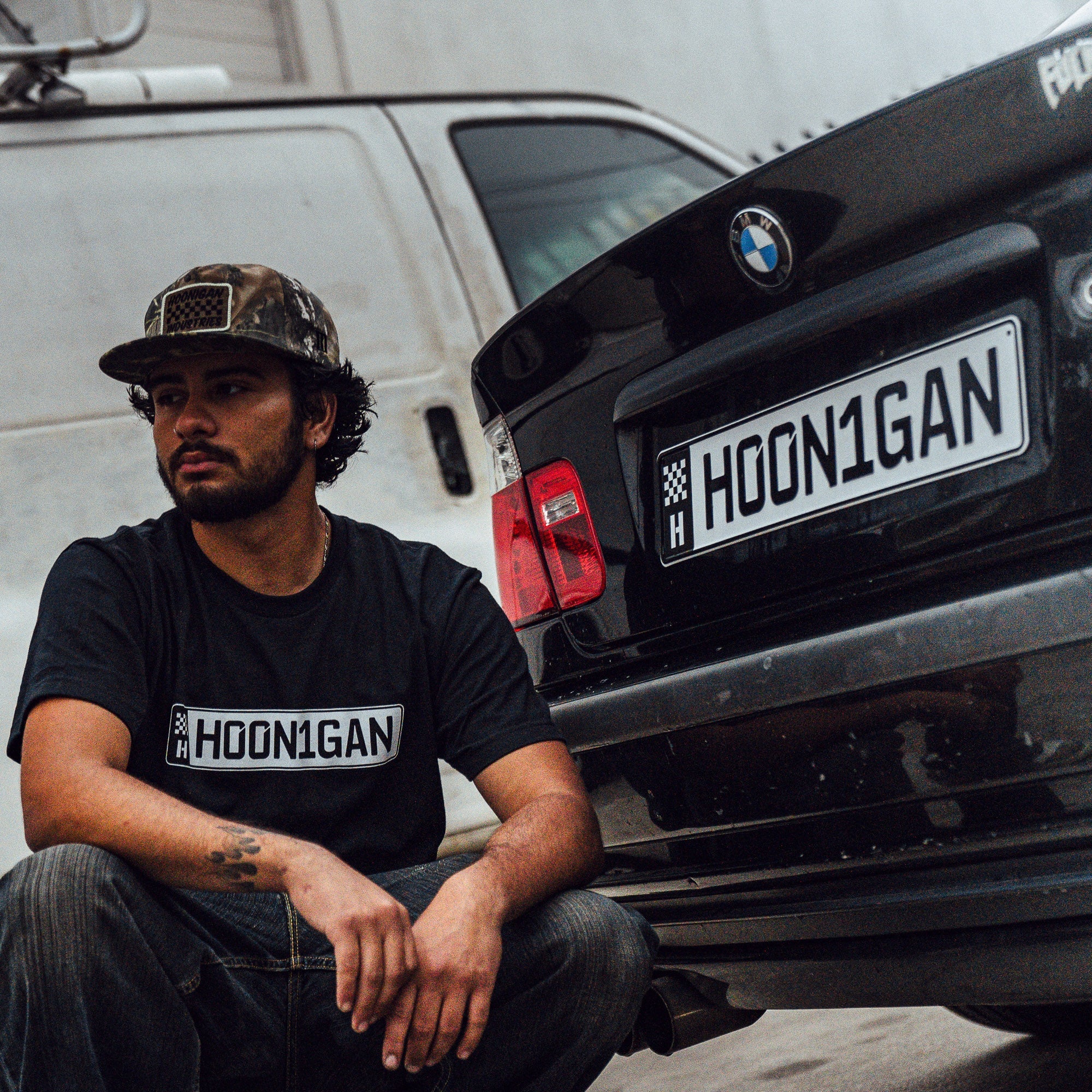 A person wearing a black Hoonigan AUTOBAHN short sleeve t-shirt sits beside a BMW, showcasing automotive culture.