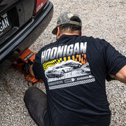A person wearing a black Hoonigan SLIDEWAYS T-shirt works on a car, showcasing a graphic of drifting cars on the shirt's back.