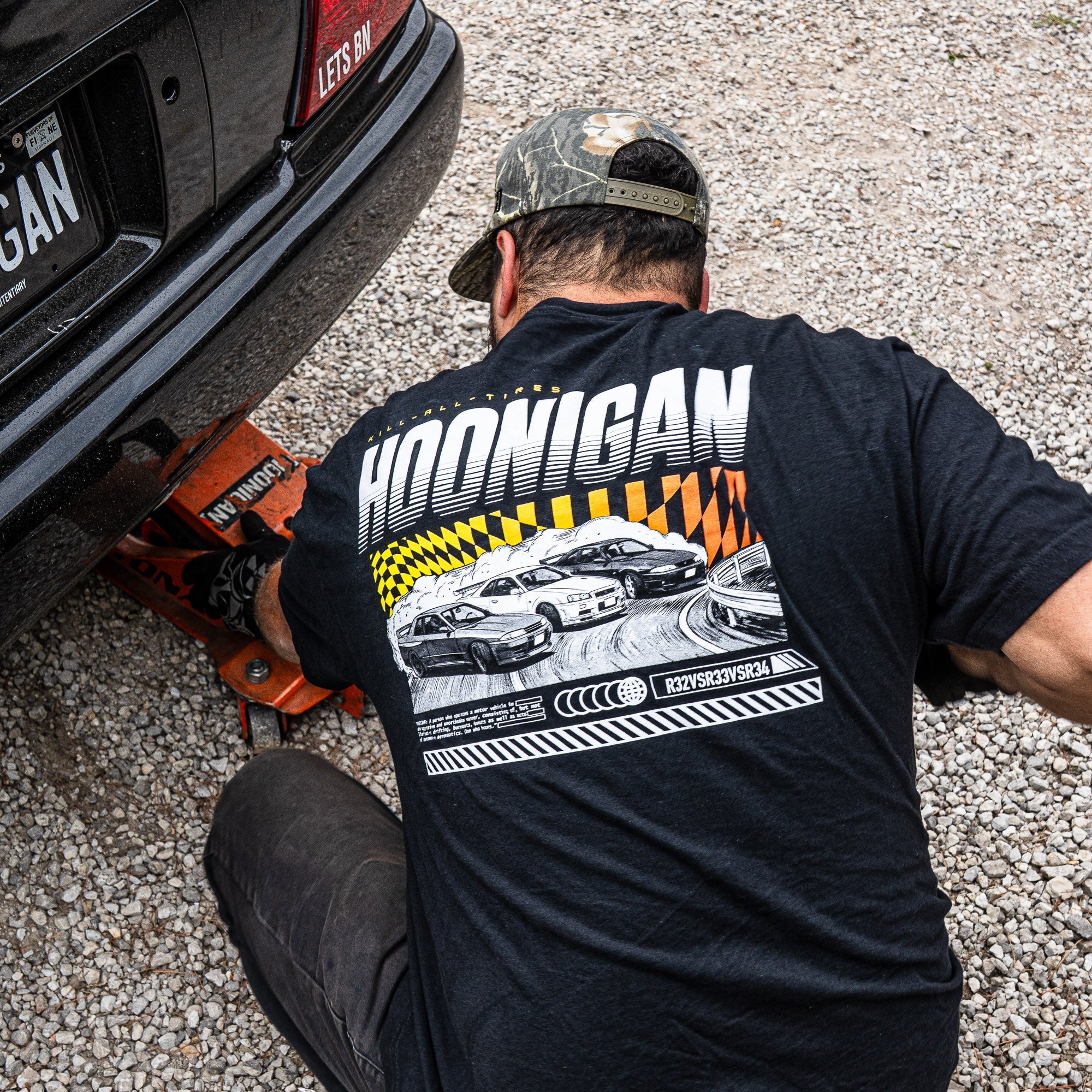 A person wearing a black Hoonigan SLIDEWAYS T-shirt works on a car, showcasing a graphic of drifting cars on the shirt's back.