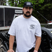 A man wearing a white Hoonigan LOW COMPRESSION short sleeve t-shirt stands next to a car, showcasing automotive lifestyle apparel.