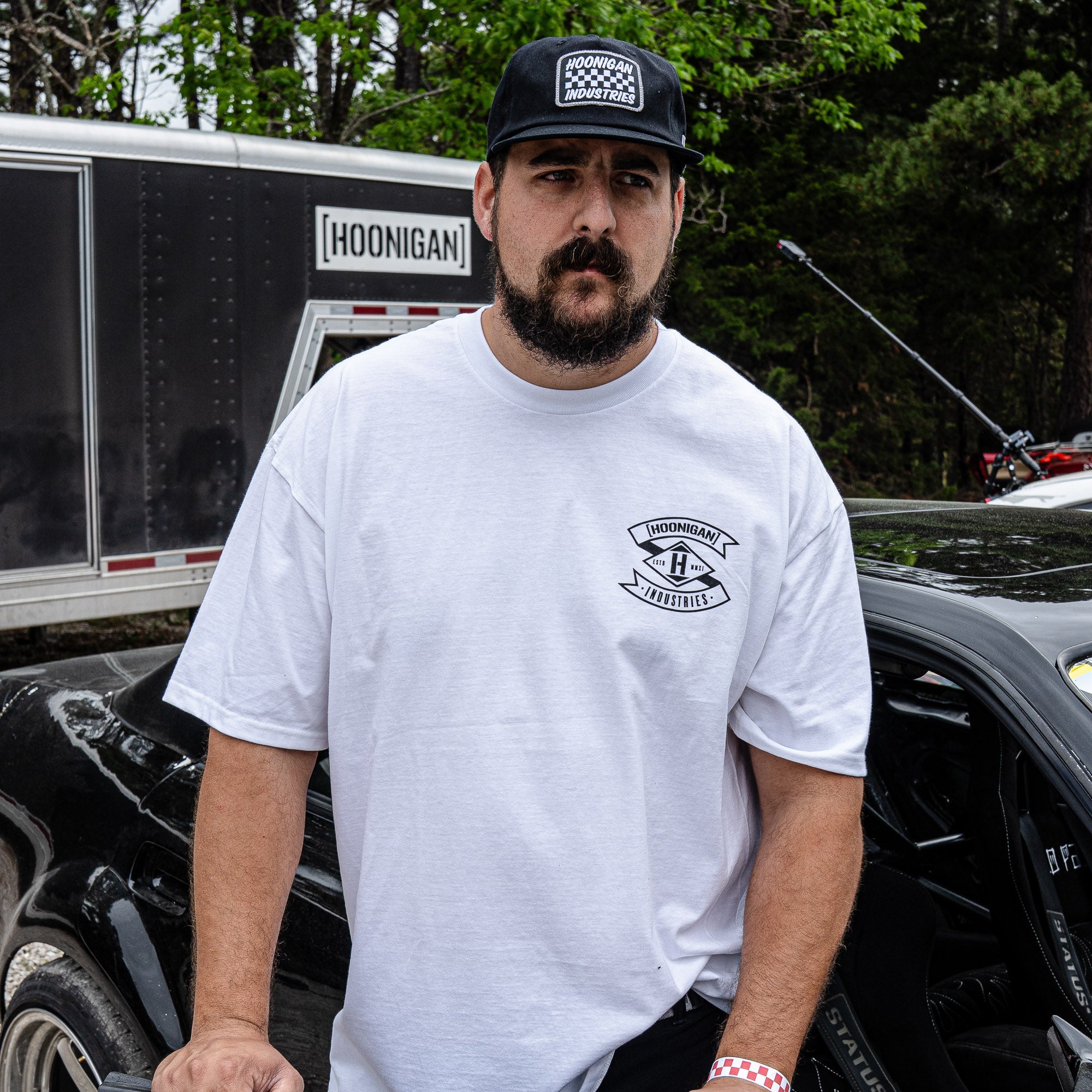 A man wearing a white Hoonigan LOW COMPRESSION short sleeve t-shirt stands next to a car, showcasing automotive lifestyle apparel.