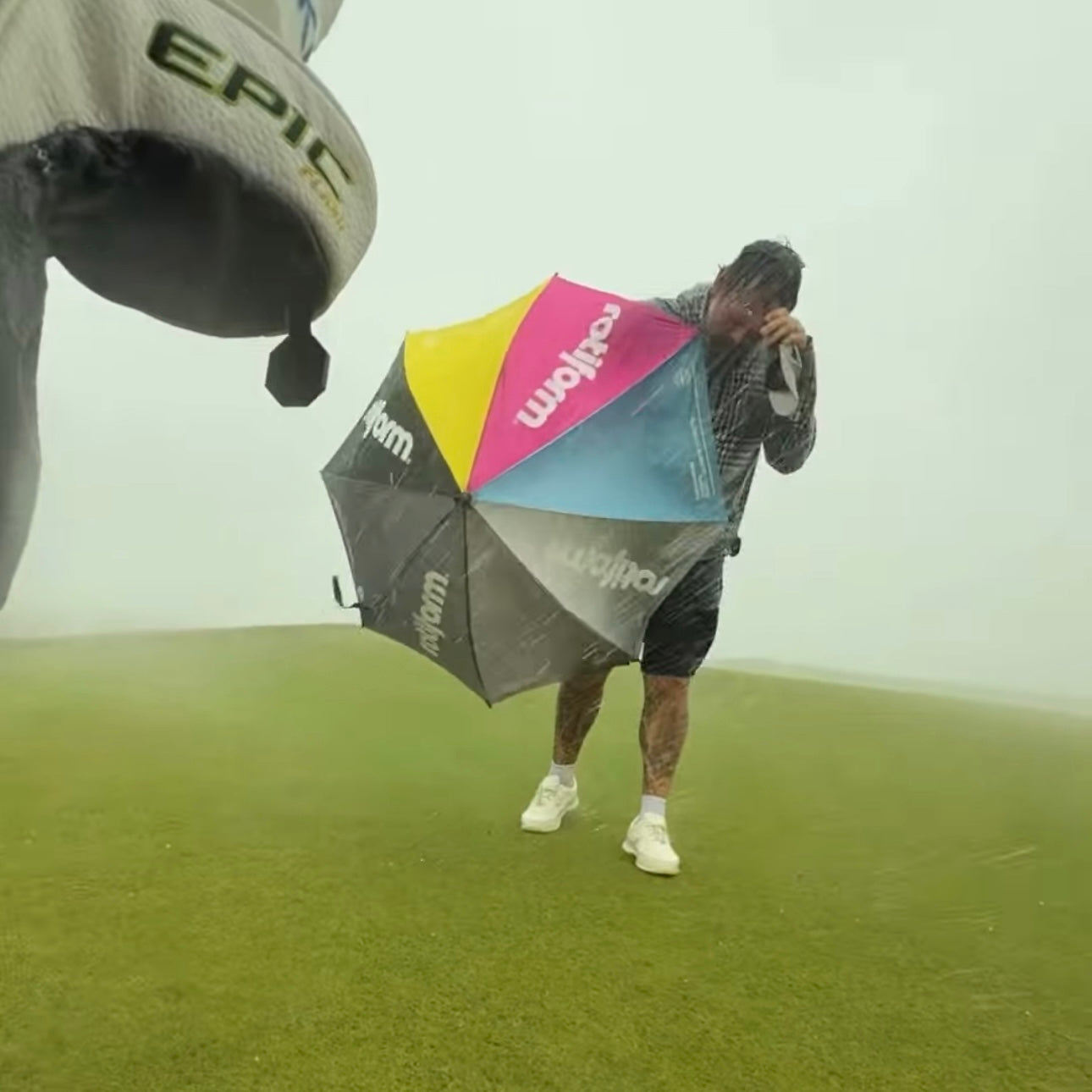 A person holding a colorful Rotiform SHADE SEEKER umbrella while standing in the rain on a grassy field.