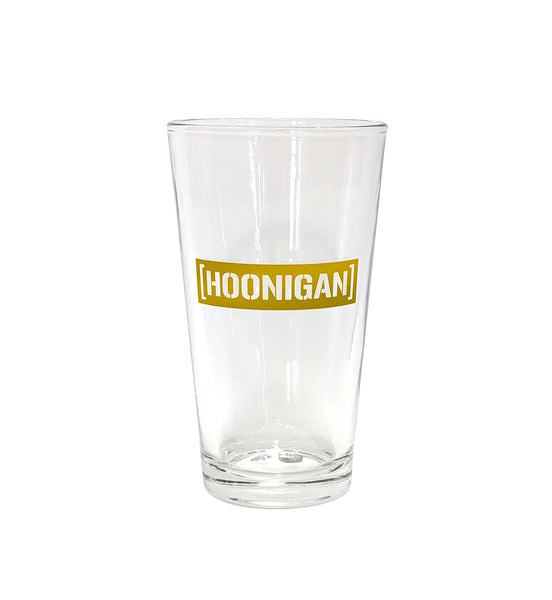 Clear pint glass featuring a bold gold Hoonigan logo, perfect for enjoying beverages while celebrating car culture.