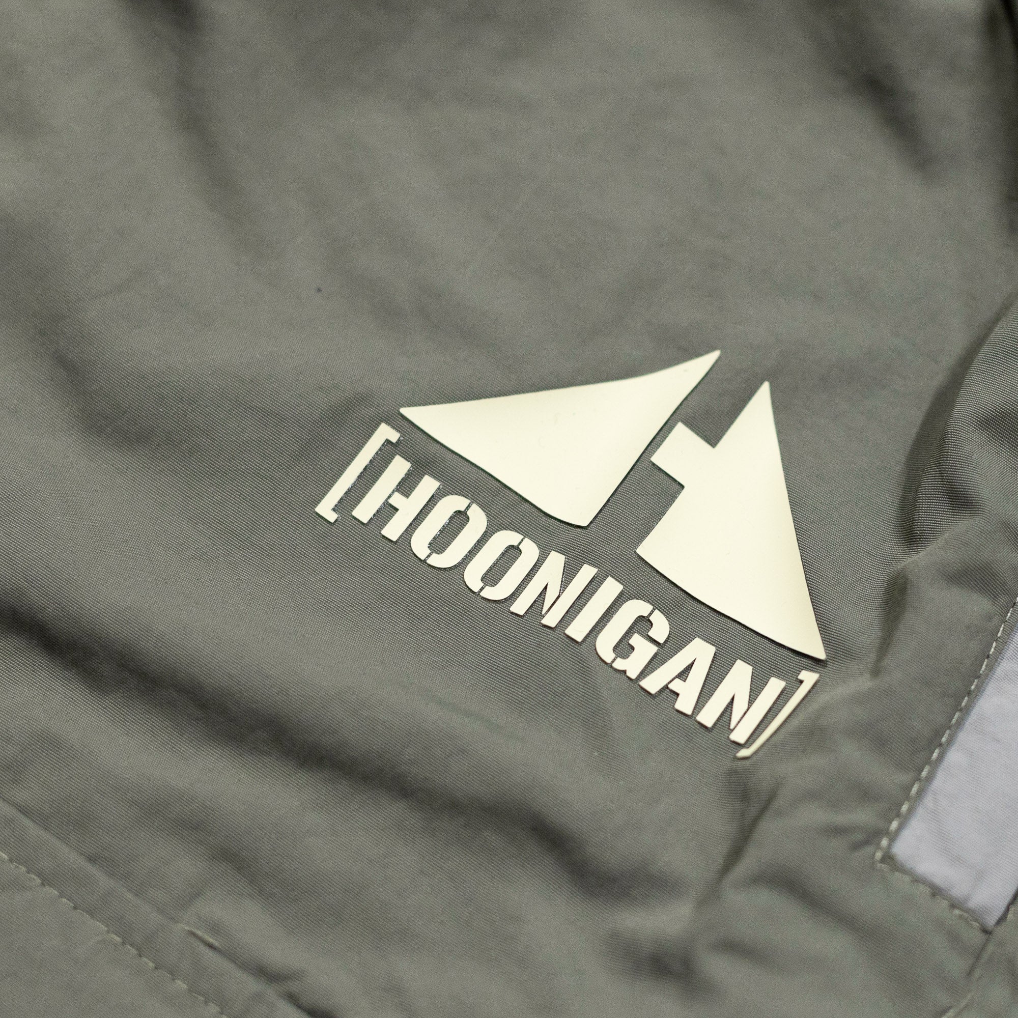 Close-up of gray cargo shorts with a rain-activated Hoonigan logo featuring two stylized triangles and bold text.