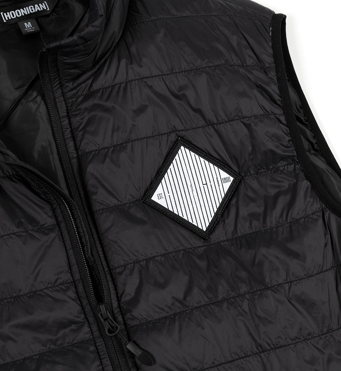 Close-up of a black GRILLE Puffer Vest with horizontal quilting, zipper closure, and a diamond-shaped patch on the chest.