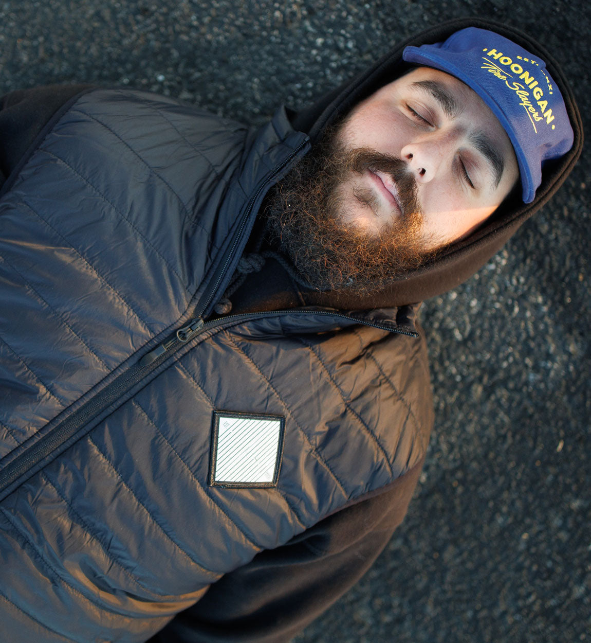 Man lying on ground wearing a black quilted puffer vest, hoodie, and a blue cap with "HONGAN" logo.