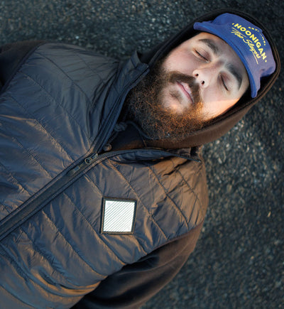 Man lying on ground wearing a black quilted puffer vest, hoodie, and a blue cap with "HONGAN" logo.