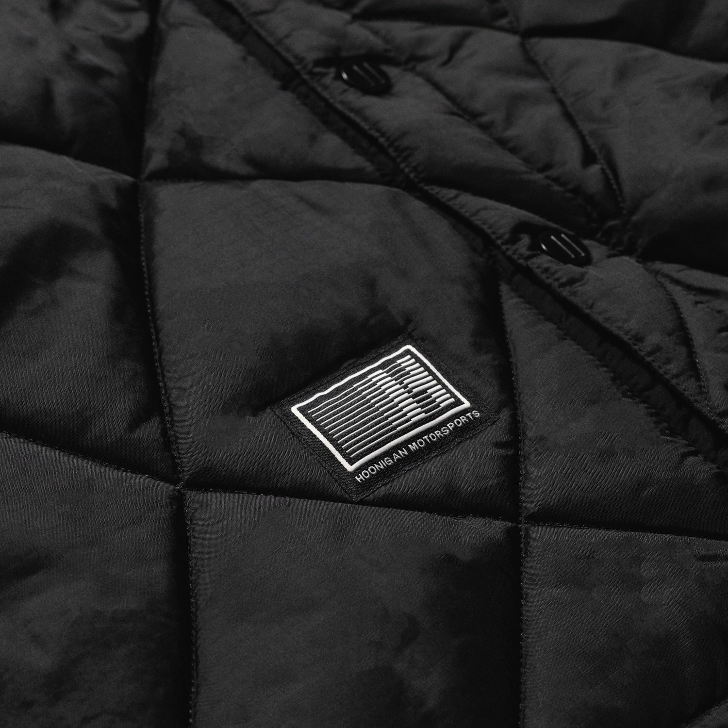 Hoonigan Motorsports VICTOR Quilted Jacket in black, featuring a zip closure, chest logo, and quilted design, displayed on a wooden background.