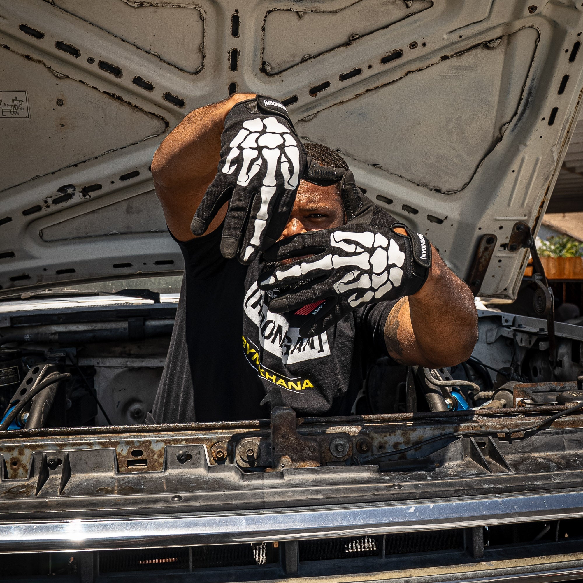 A person wearing Hoonigan FIND OUT Gloves poses with hands shaped like a skeleton while working on a car engine.