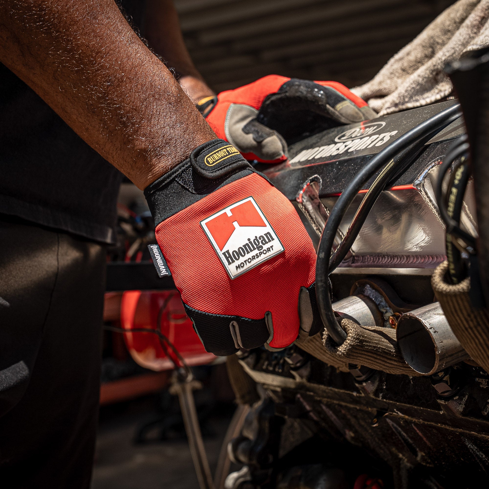 Hoonigan Surgeon General gloves worn by a mechanic working on a car engine in a garage setting.