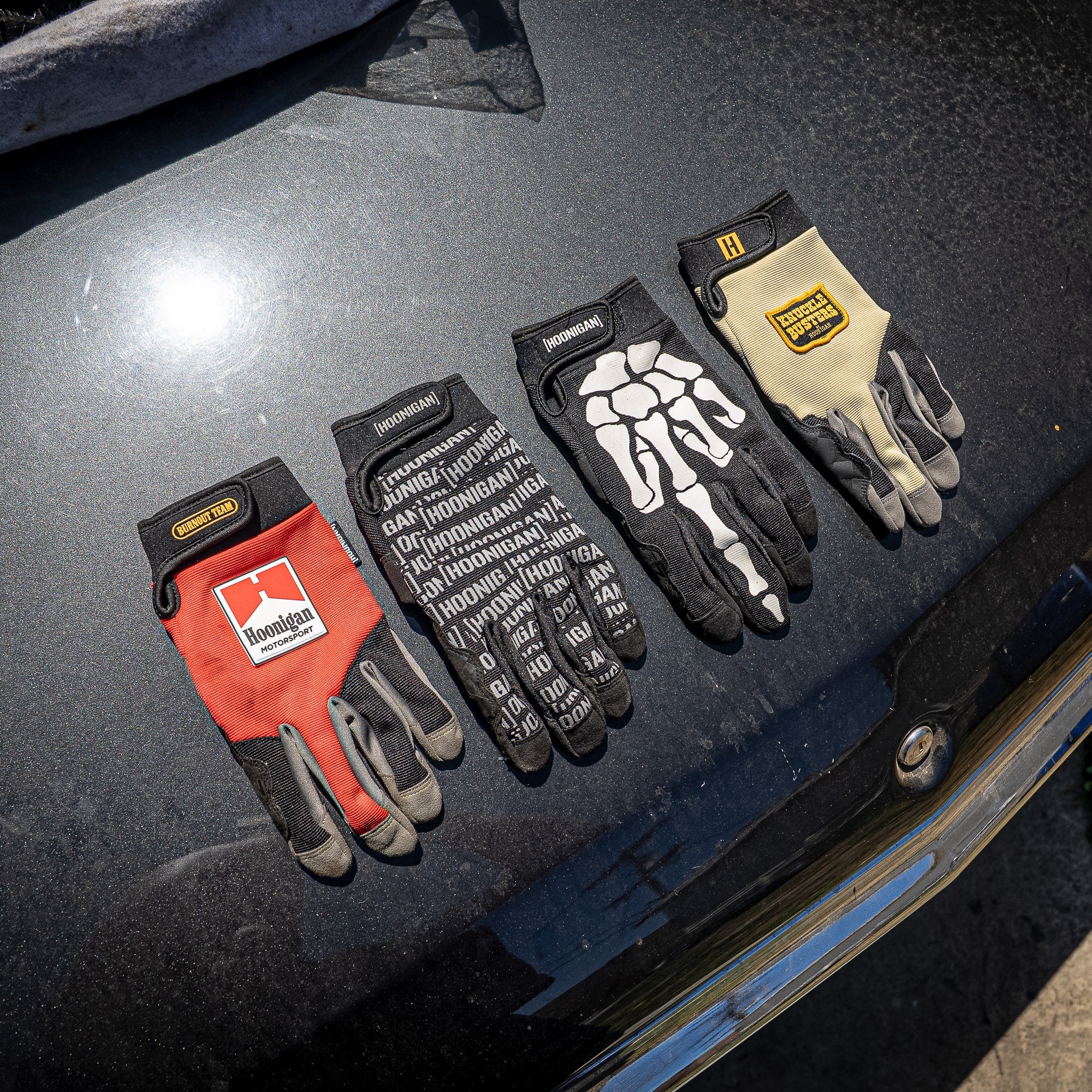 Hoonigan FIND OUT Gloves displayed on a car hood, featuring various designs for motorsport enthusiasts.