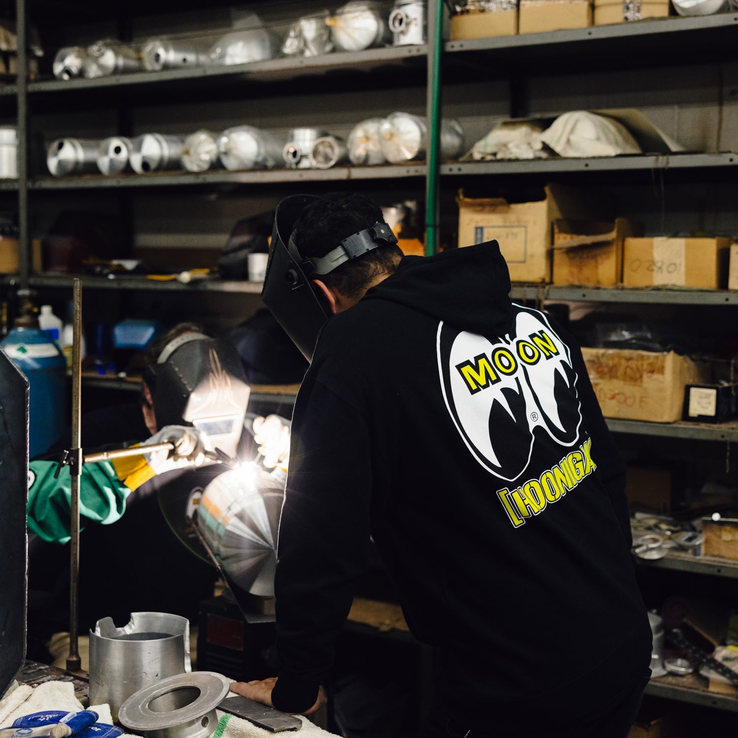 Hoonigan x Mooneyes pullover hoodie worn by a person working on automotive parts in a workshop.
