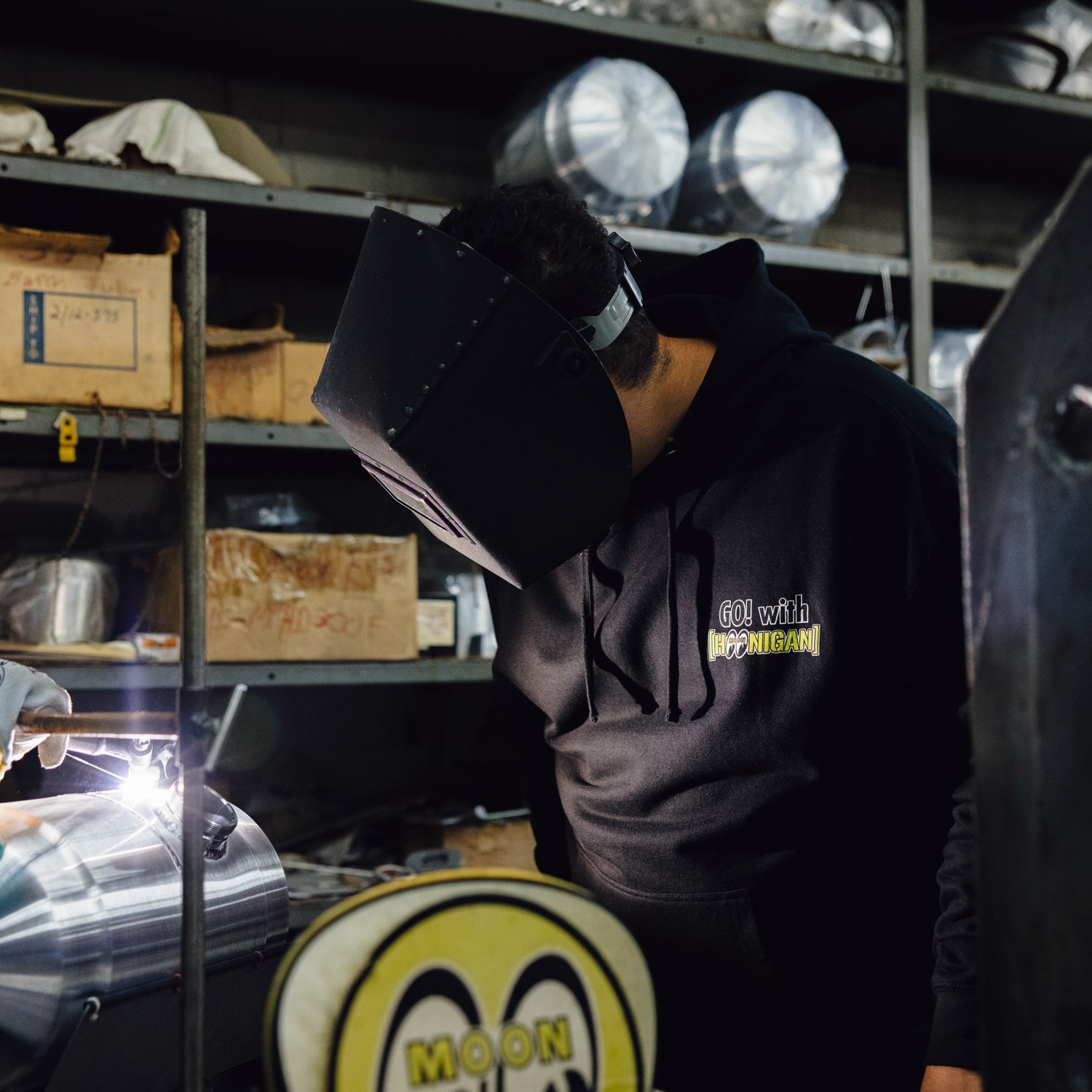A person wearing a Hoonigan x Mooneyes pullover hoodie works on metal fabrication, focused on welding in a workshop.