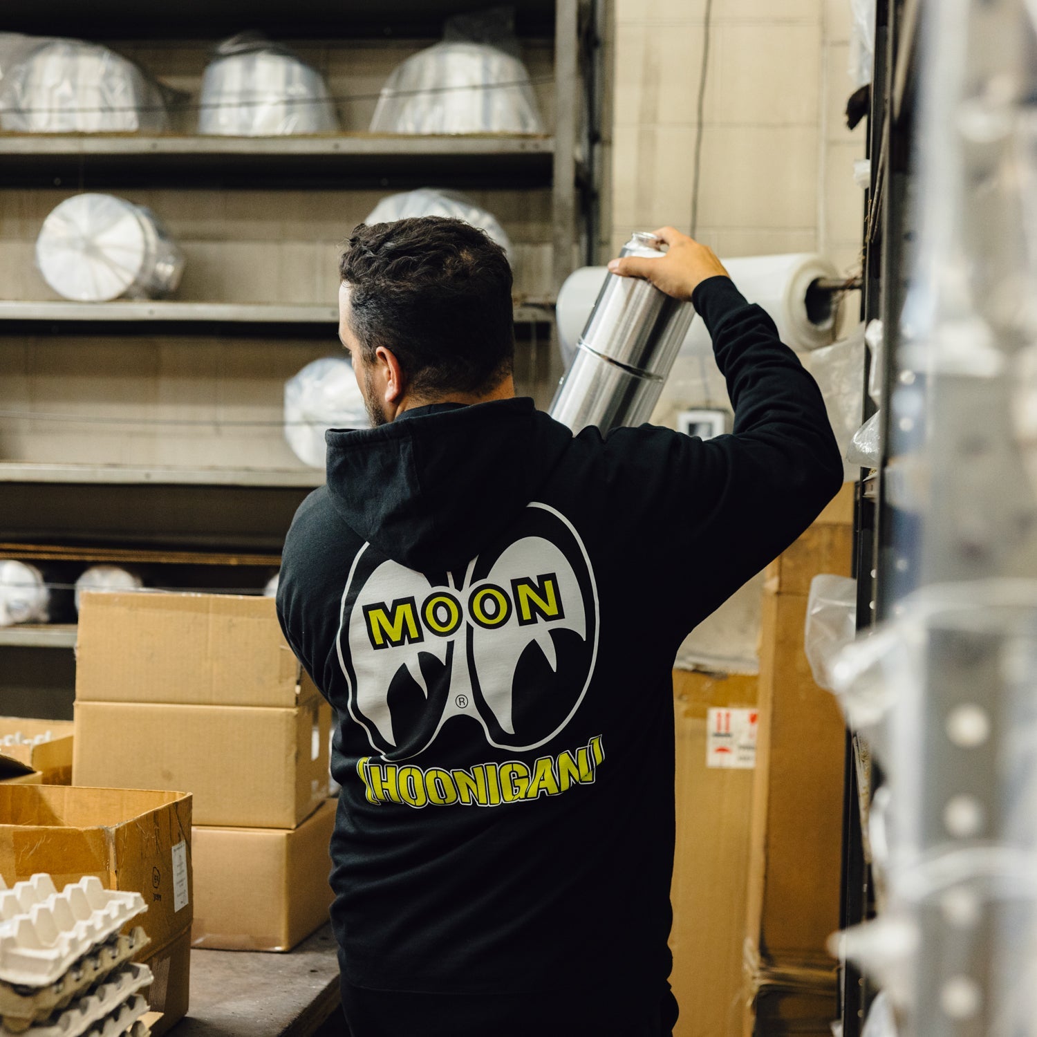Man wearing a Hoonigan x Mooneyes pullover hoodie, shaking a metal canister in a warehouse setting.
