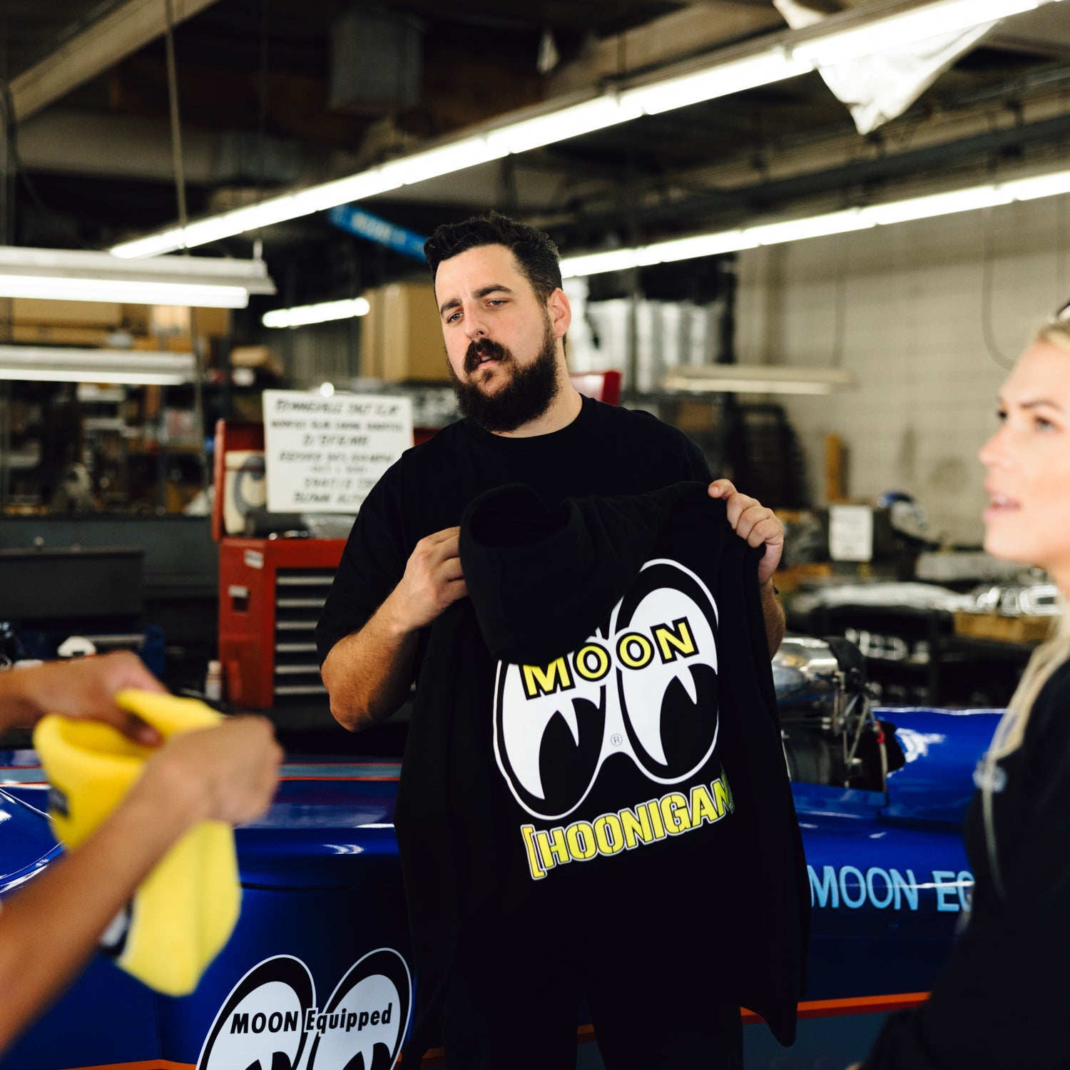 A man holds a Hoonigan x Mooneyes pullover hoodie featuring bold graphics, surrounded by automotive enthusiasts in a workshop.