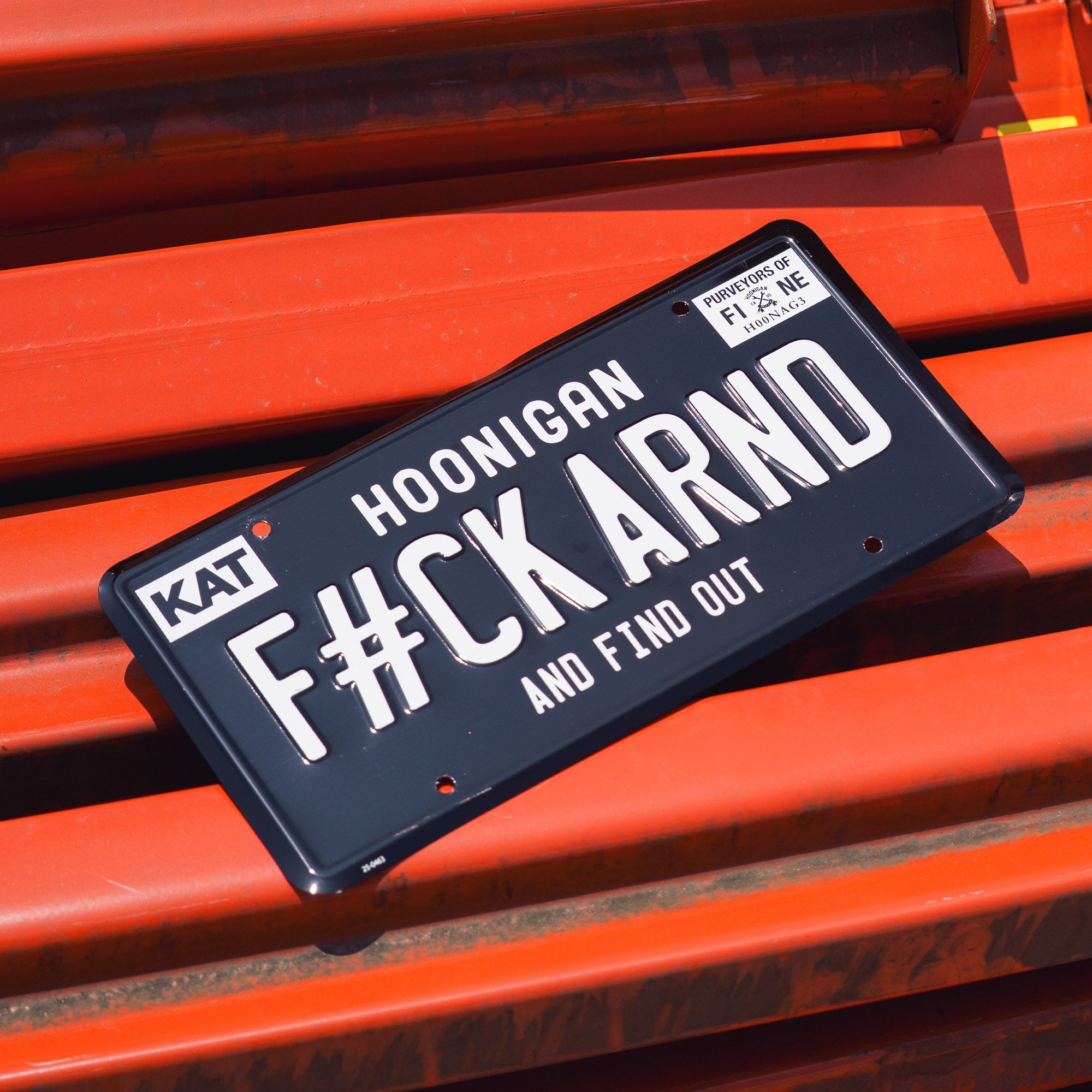 Black license plate with white text reading "FCK ARND" and "and find out," mounted on an orange surface.