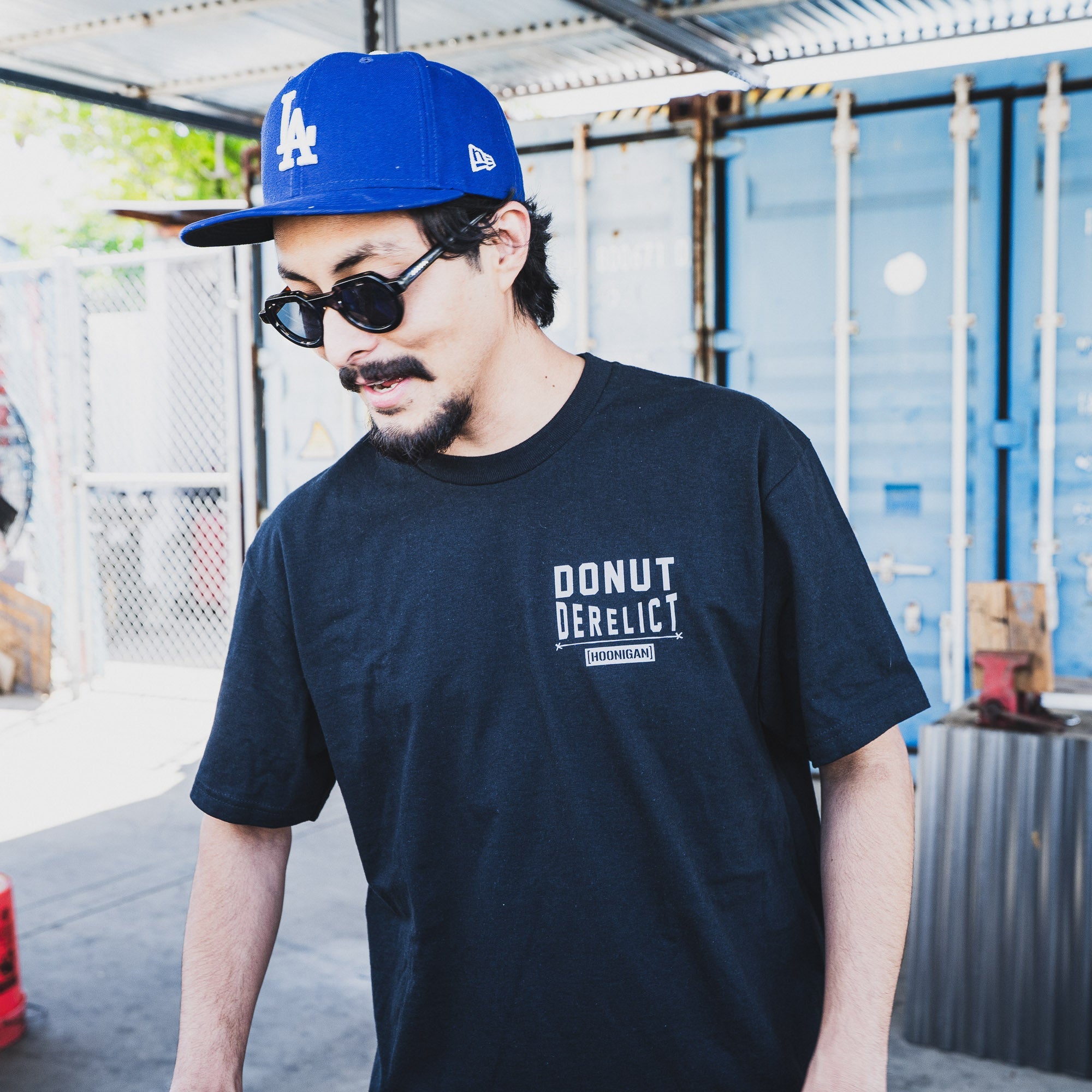 Man wearing a black Hoonigan DONUT DERELICT short sleeve t-shirt, sunglasses, and a blue cap in an industrial setting.