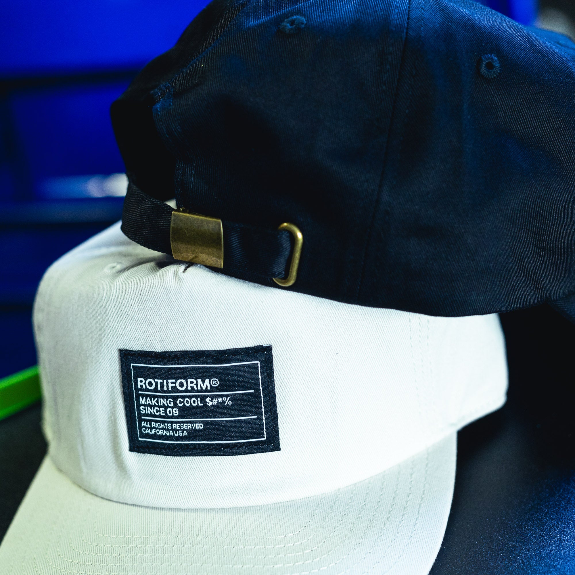 Black and white unstructured strapback hats with "Making Cool S#*% Since 09" label, featuring adjustable strap and metal buckle.
