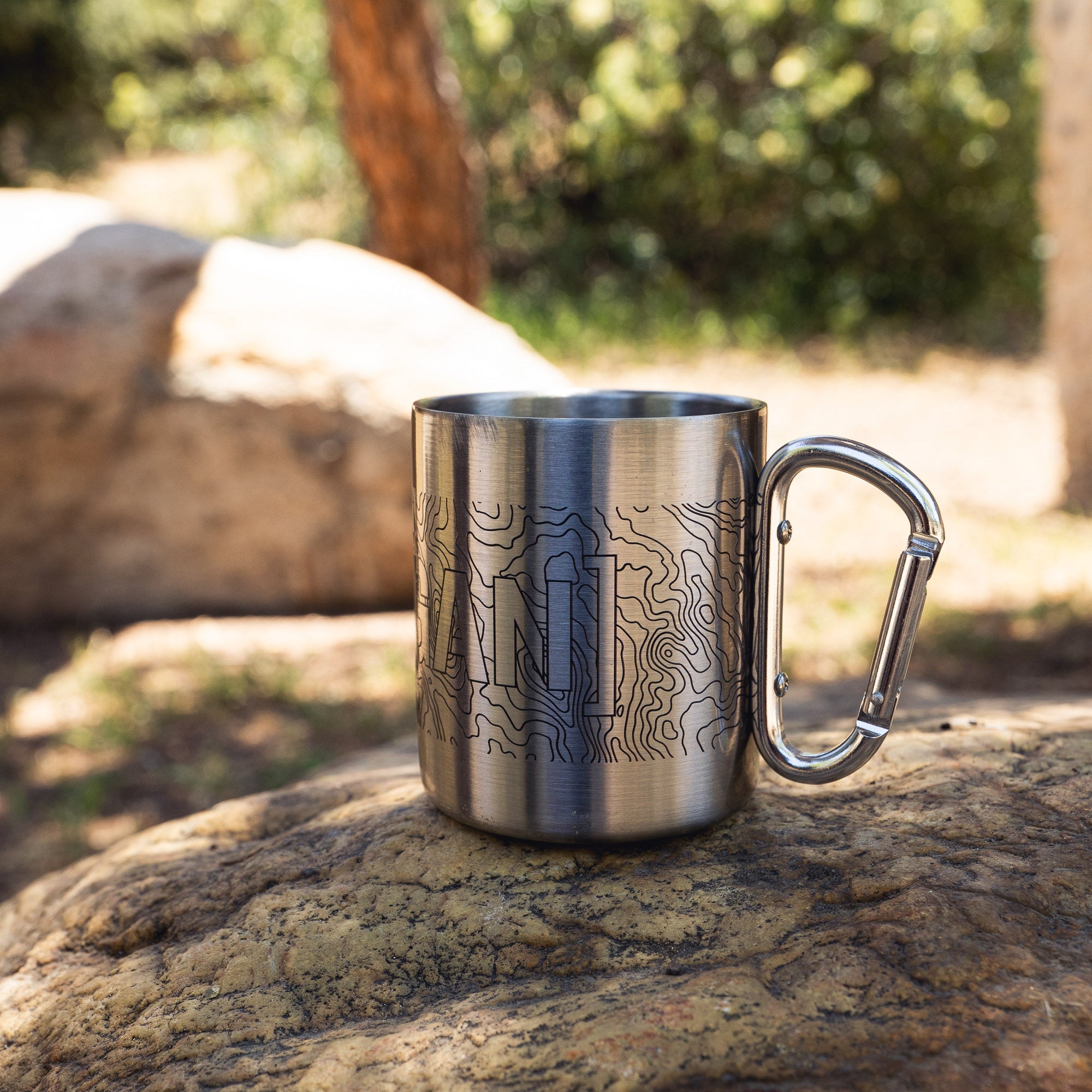 Stainless steel mug with a carabiner handle featuring topographic map design and the Hoonigan logo, ideal for outdoor use.