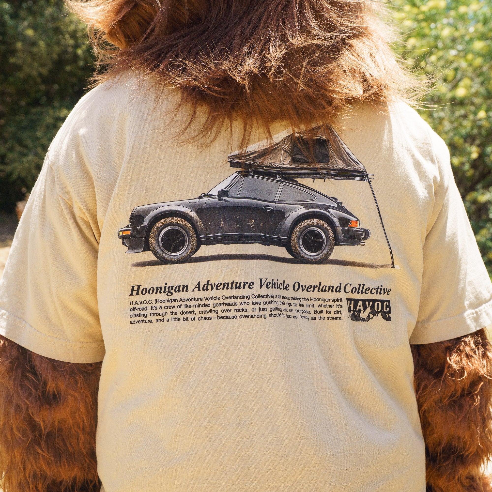 Close-up of a person wearing a beige Hoonigan SAFARI Short Sleeve Tee featuring a black vintage car with a roof tent and overland graphic design.