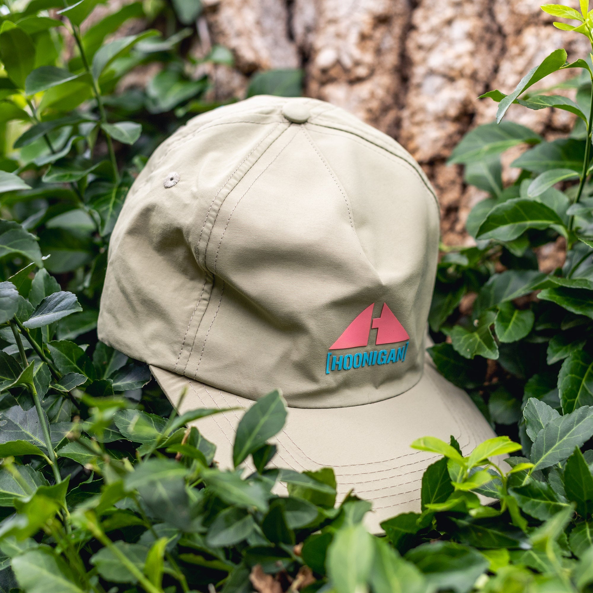 Beige Hoonigan H.A.V.O.C. unstructured strapback hat with colorful logo, resting among green leaves outdoors.