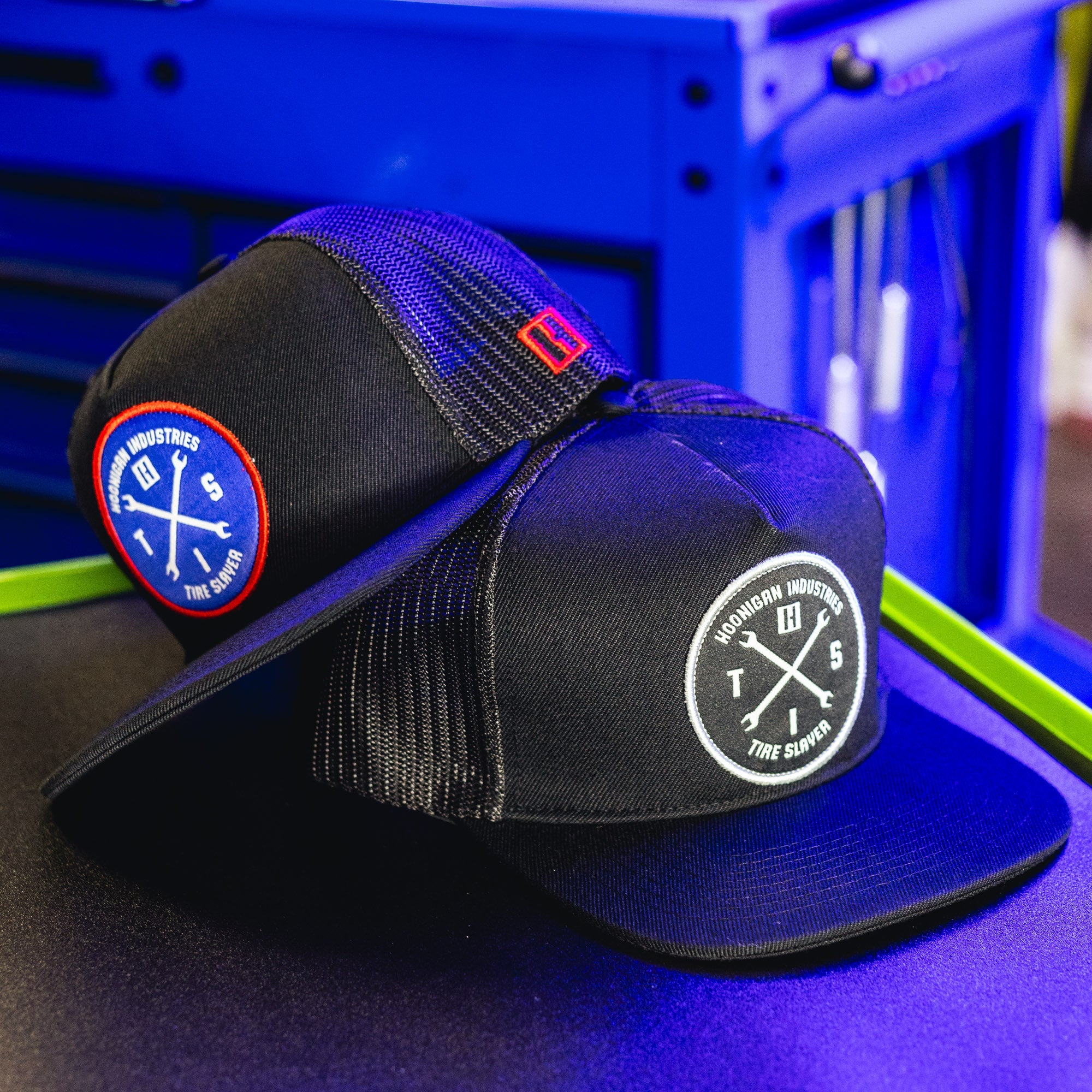 Black trucker hat with Hoonigan Industries logo patch, mesh back, and curved brim, featuring a circular patch with crossed tools.