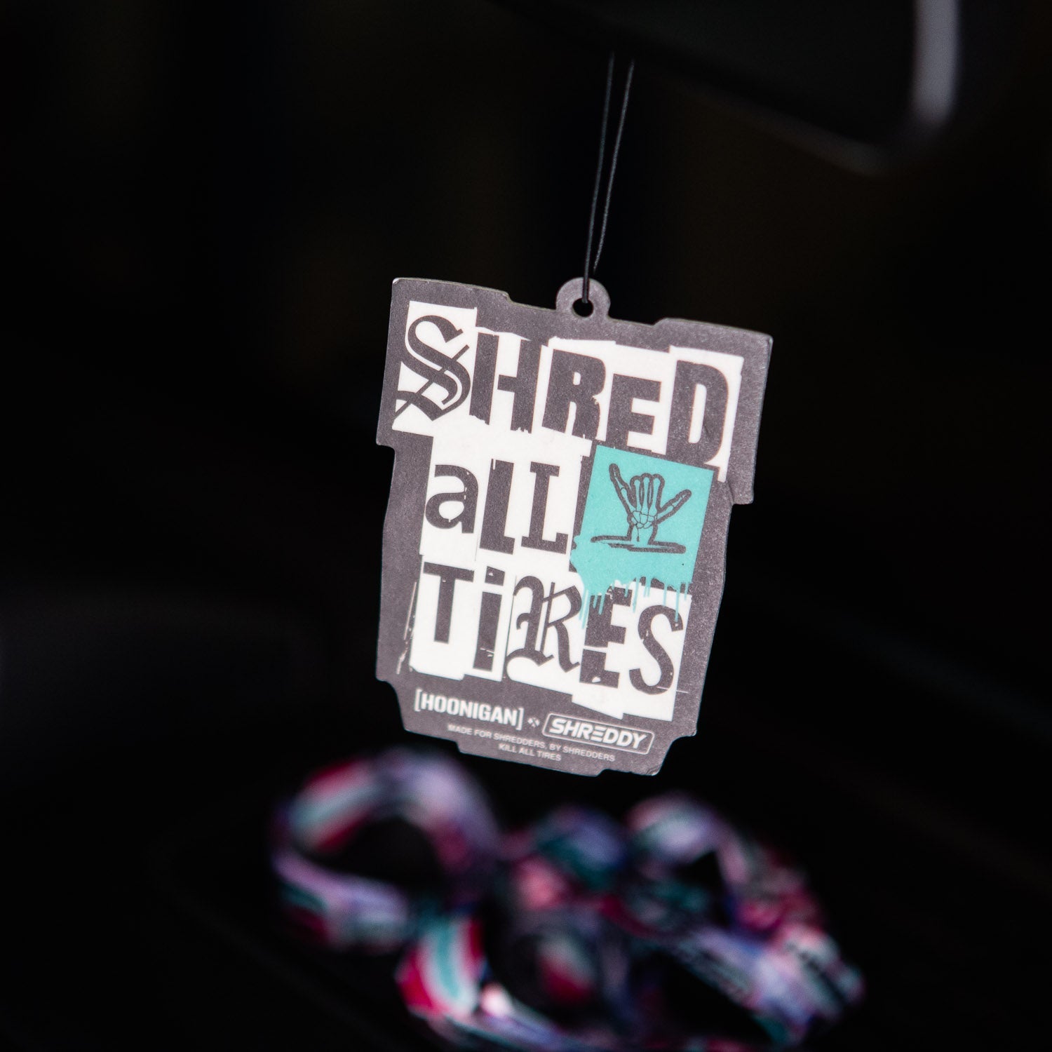 Hoonigan x Shreddy SHRED ALL TIRES air freshener hanging in a car, featuring bold graphics and vibrant colors.