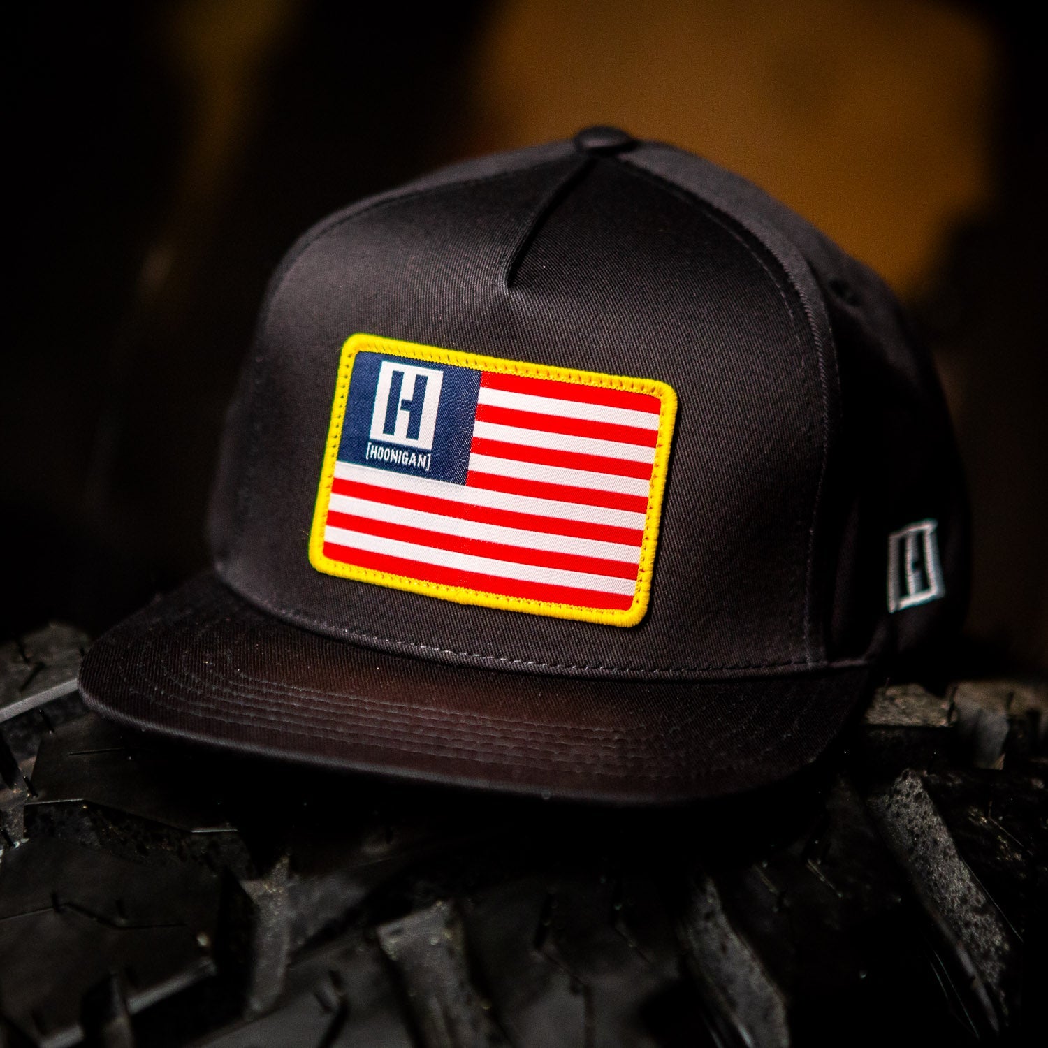 Black snapback hat featuring a patriotic American flag patch with the Hoonigan logo, perfect for motorsport enthusiasts.