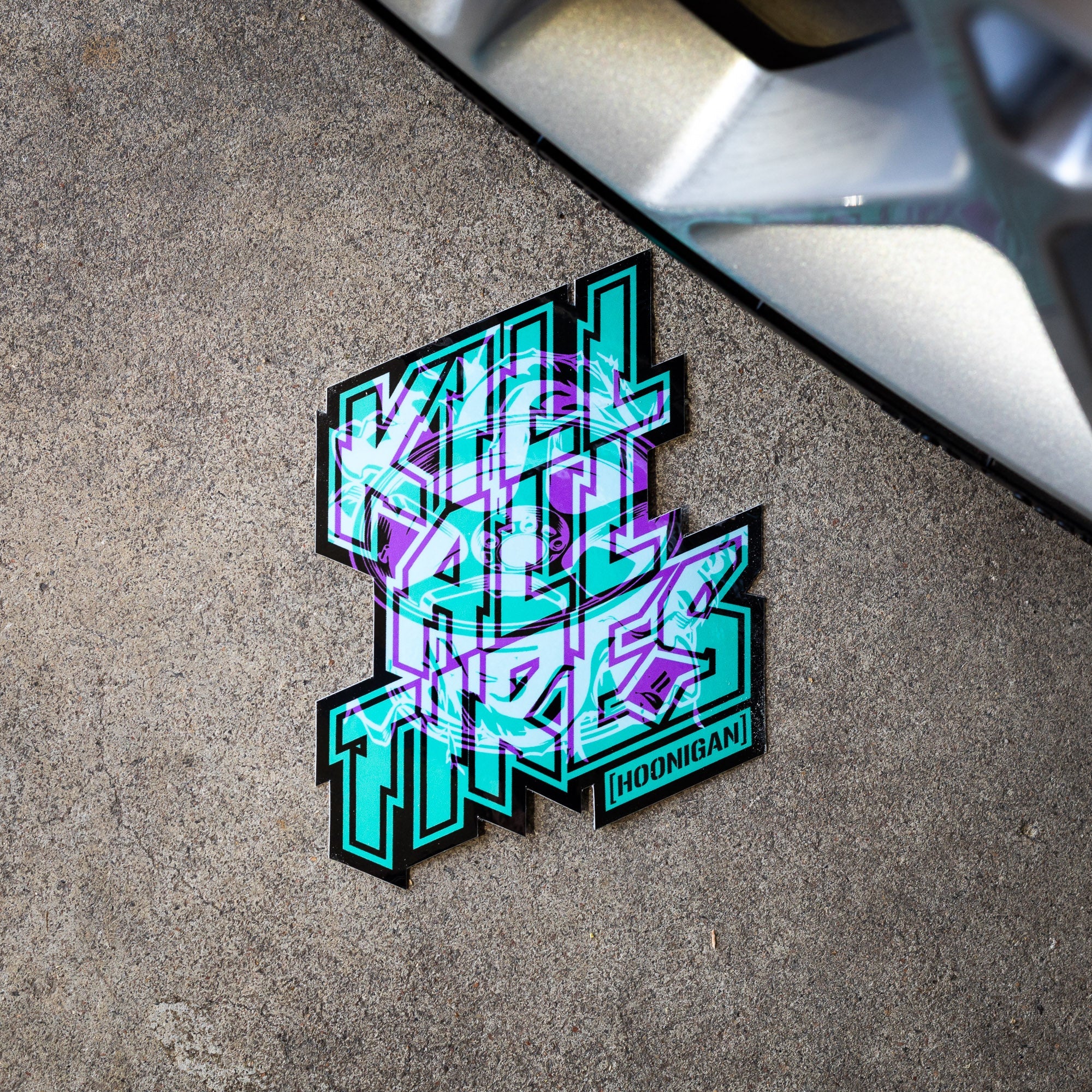 Hoonigan TREADWEAR KILL ALL TIRES sticker featuring bold, colorful text on a concrete surface near a wheel.