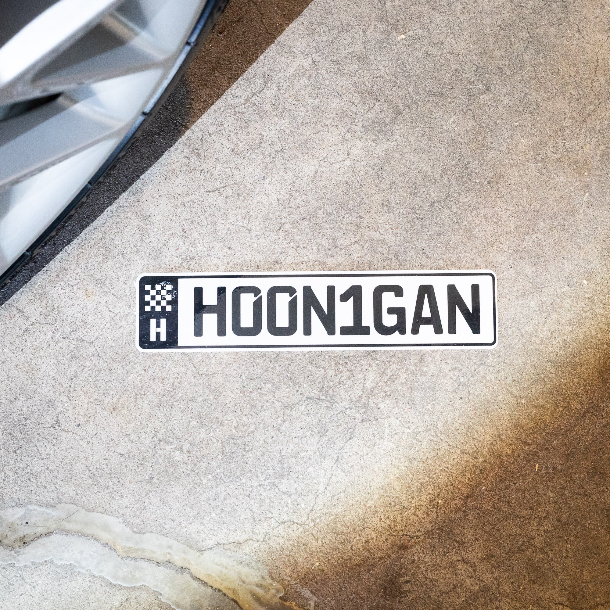 Hoonigan AUTOBAHN sticker featuring bold lettering on a concrete surface near a car wheel. Ideal for automotive enthusiasts.