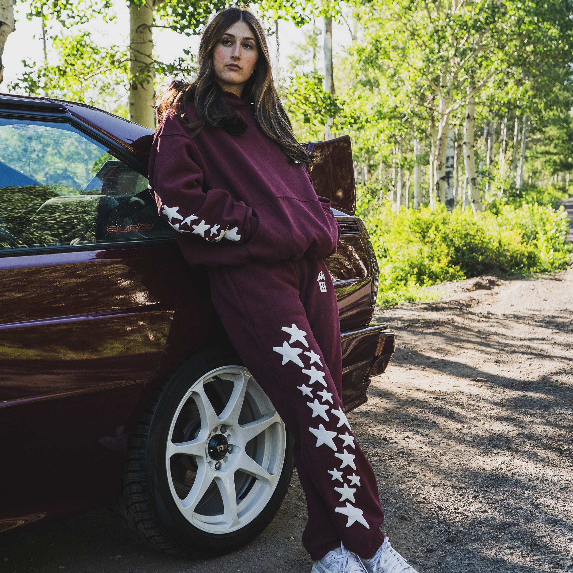 A person leans against a car in a natural setting, wearing Hoonigan x Lia Block UR STAR Sweatpants featuring star designs.