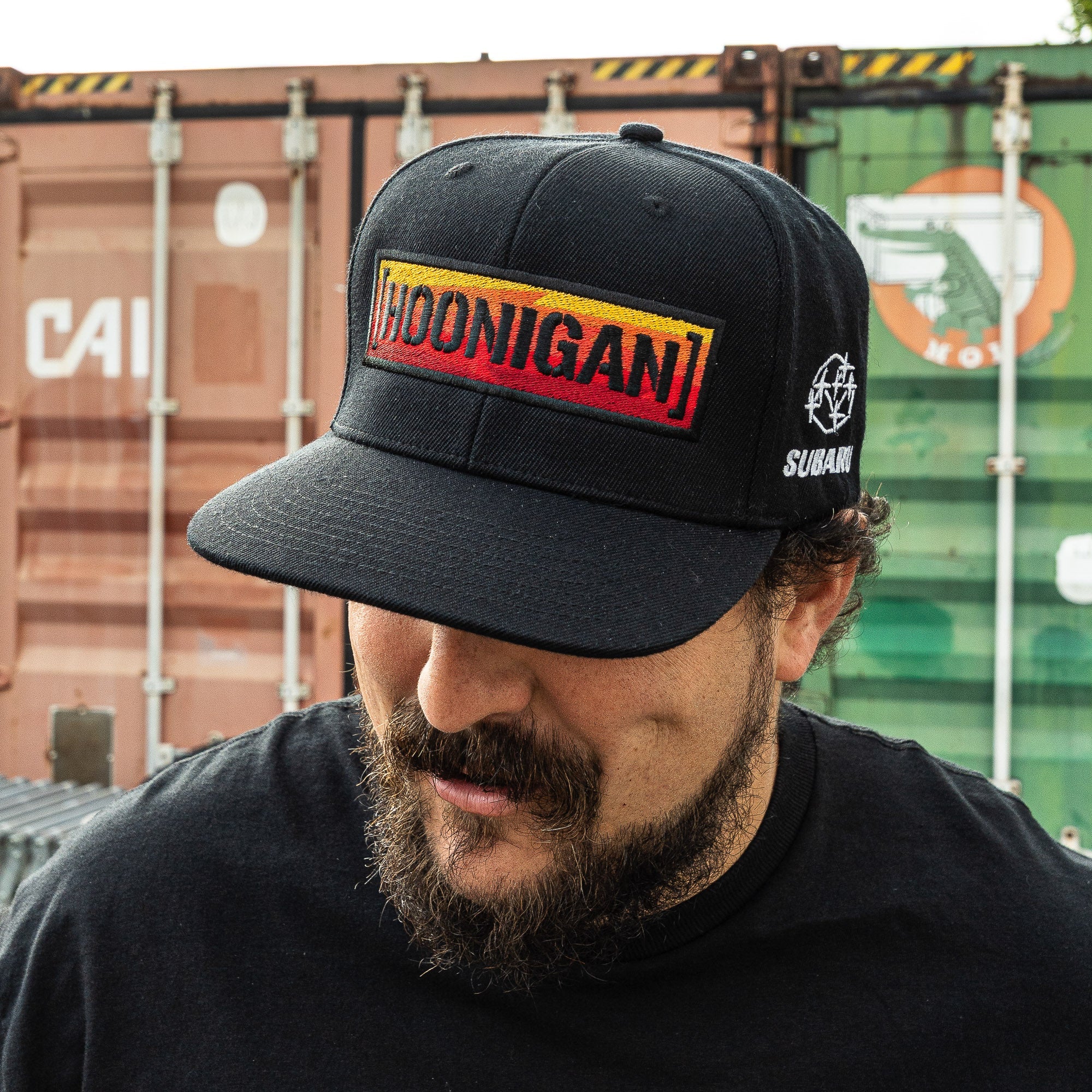 Black snapback hat featuring the Hoonigan logo in a bold gradient design, worn by a man with a beard.