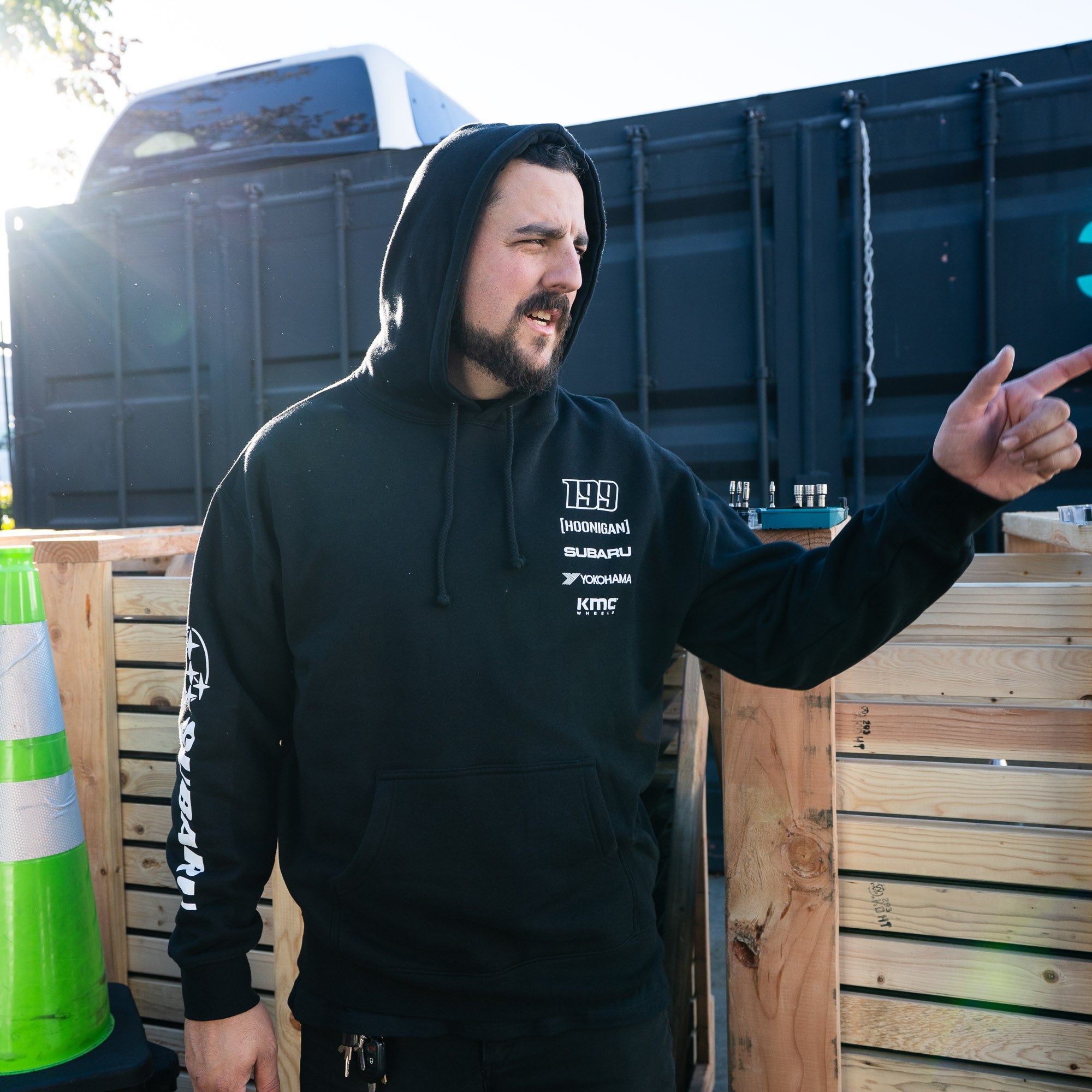 Hoonigan GYMKHANA AUSSIE SHRED LIVERY Pullover Hoodie worn by a man gesturing outdoors, showcasing automotive lifestyle style.