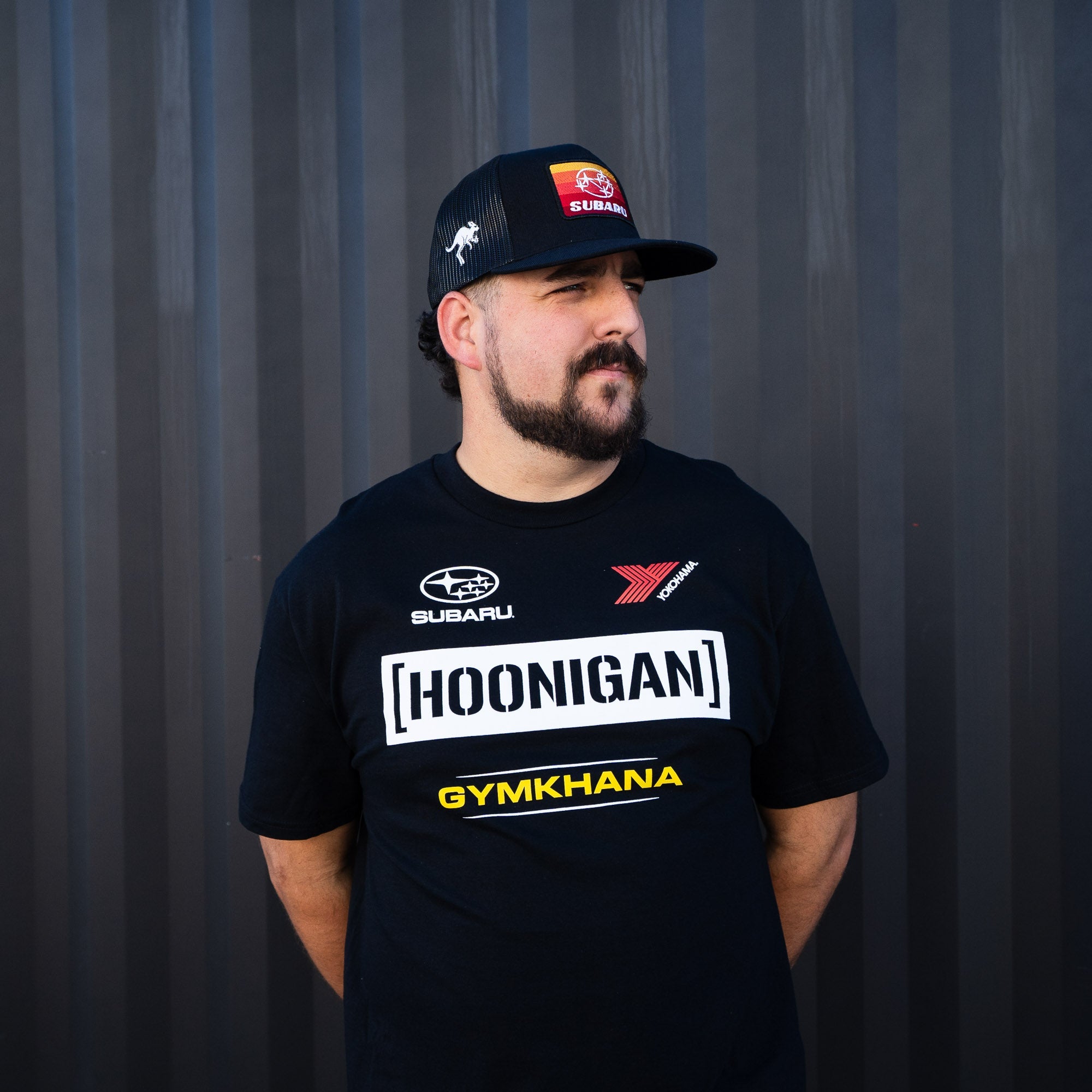 Man wearing a black Hoonigan GYMKHANA AUSSIE SHRED HORIZON trucker snapback hat and a matching black T-shirt.