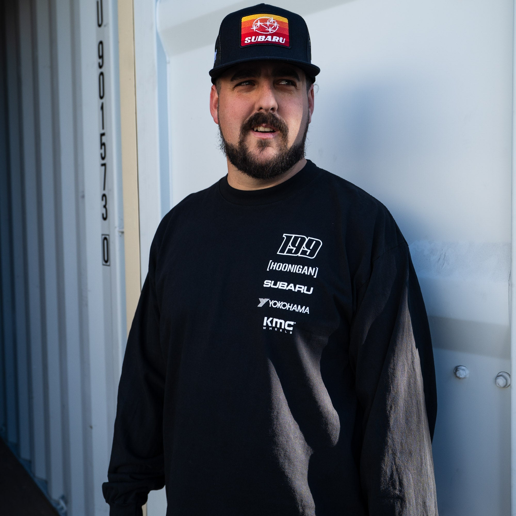 Man wearing a Hoonigan GYMKHANA AUSSIE SHRED HORIZON Trucker Snapback Hat and a long-sleeve shirt, standing against a container.