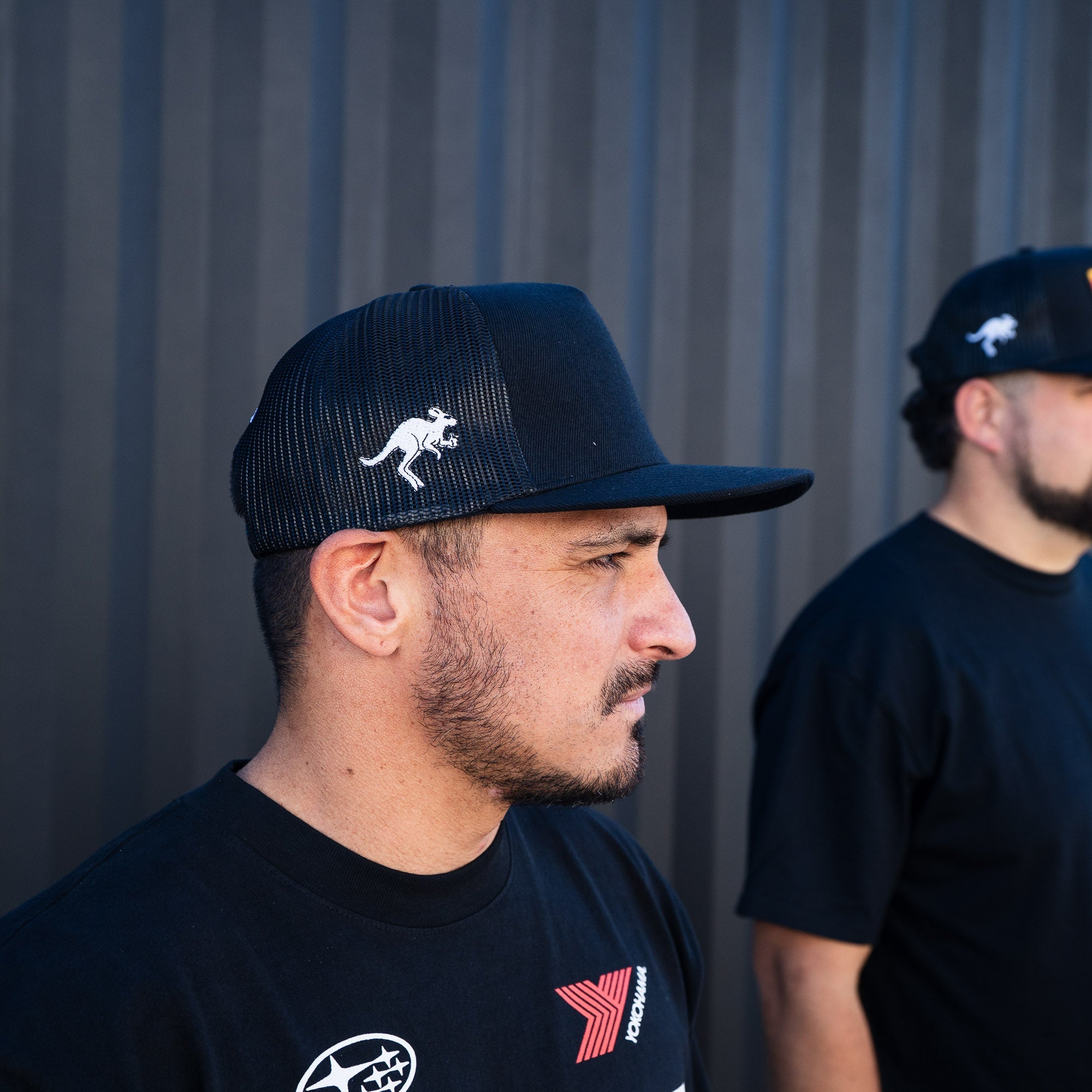 Black trucker strapback hat featuring a kangaroo logo, worn by a man alongside another individual in casual attire.