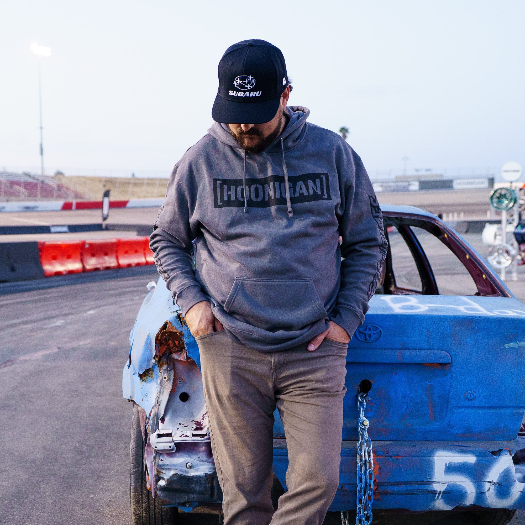 Hoonigan CENSOR BAR KILL ALL TIRES pullover hoodie worn by a man standing next to a damaged car at a motorsport venue.
