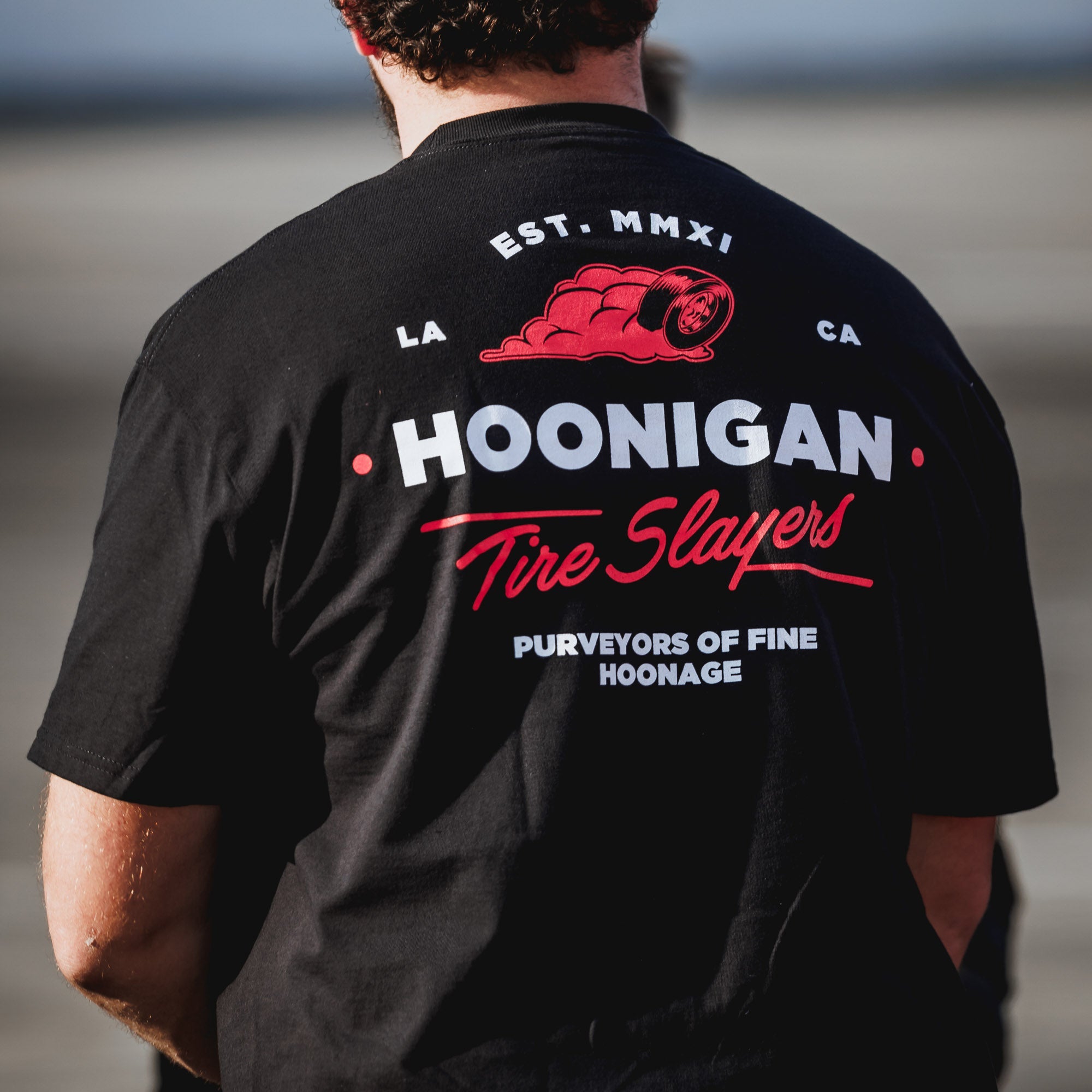Black short sleeve t-shirt featuring Hoonigan logo and "Tire Slayers" text on the back, perfect for motorsport enthusiasts.