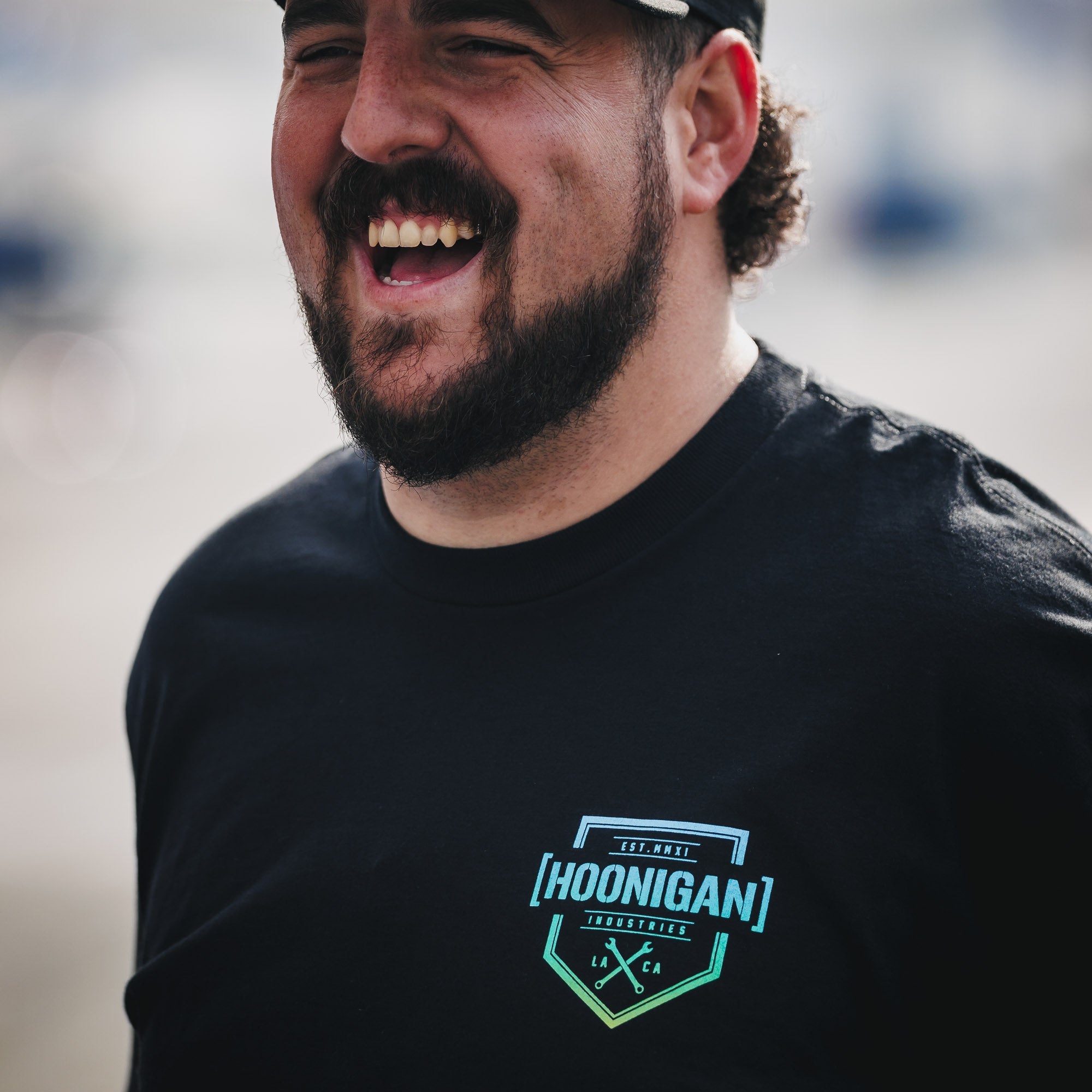 Man smiling while wearing a black Hoonigan BRACKET X LOGO short sleeve t-shirt, showcasing automotive lifestyle apparel.