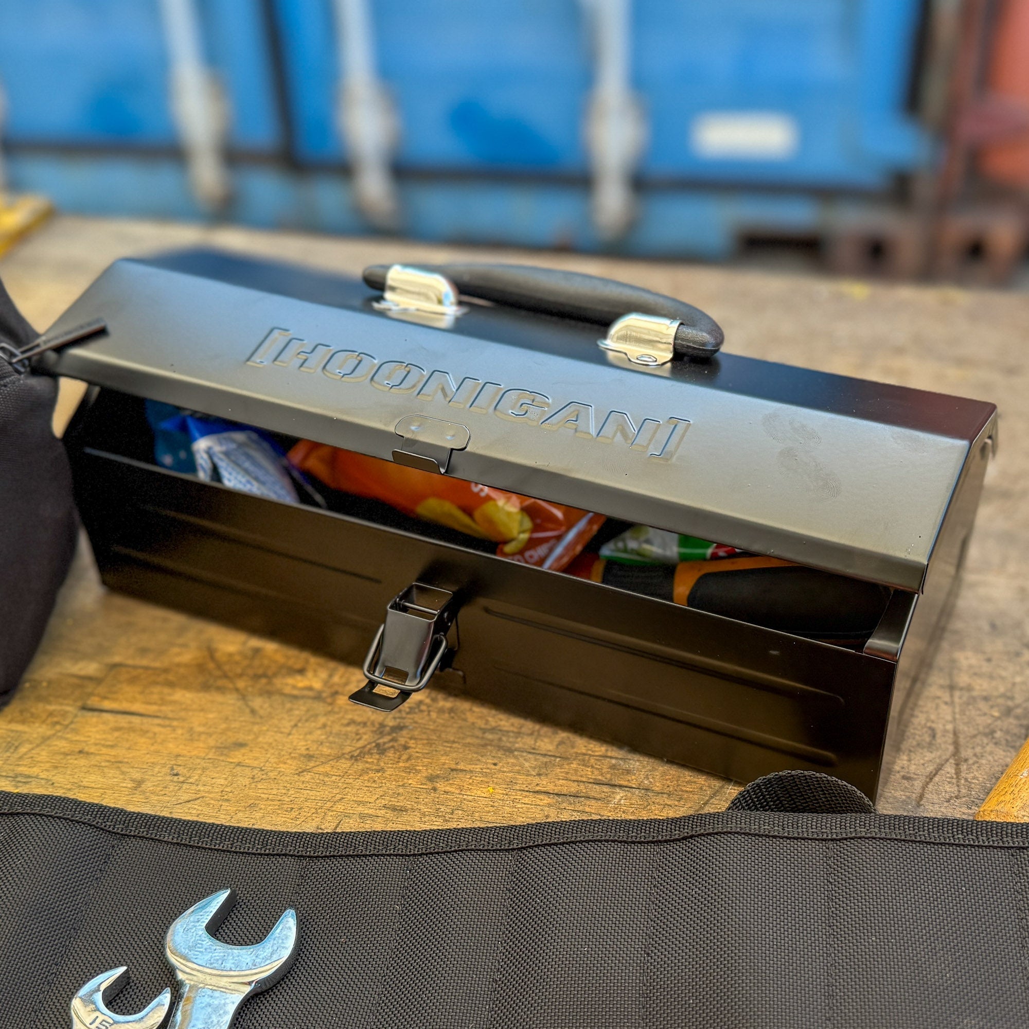 Hoonigan metal tool box open on a workbench, revealing tools and supplies inside, with a wrench beside it.