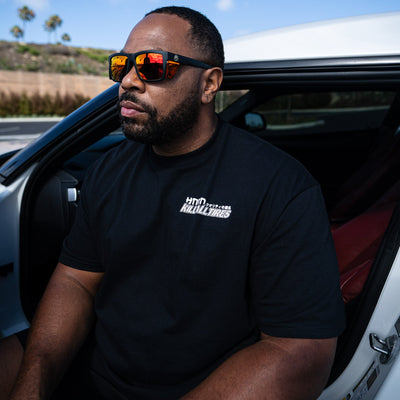 Man wearing a black Hoonigan WANGAN C1 short sleeve tee and sunglasses, sitting in a car with a sporty interior.