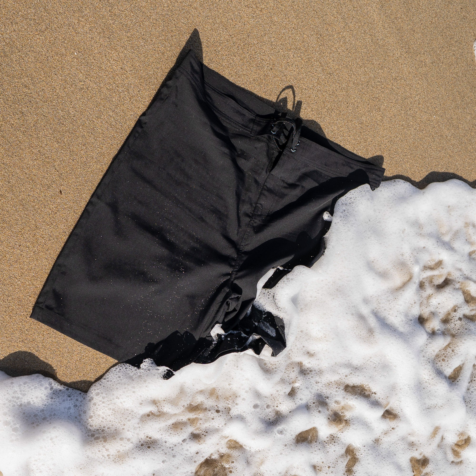 Hoonigan OIL SLICK Board Shorts laid on sandy beach with ocean waves gently washing over them.