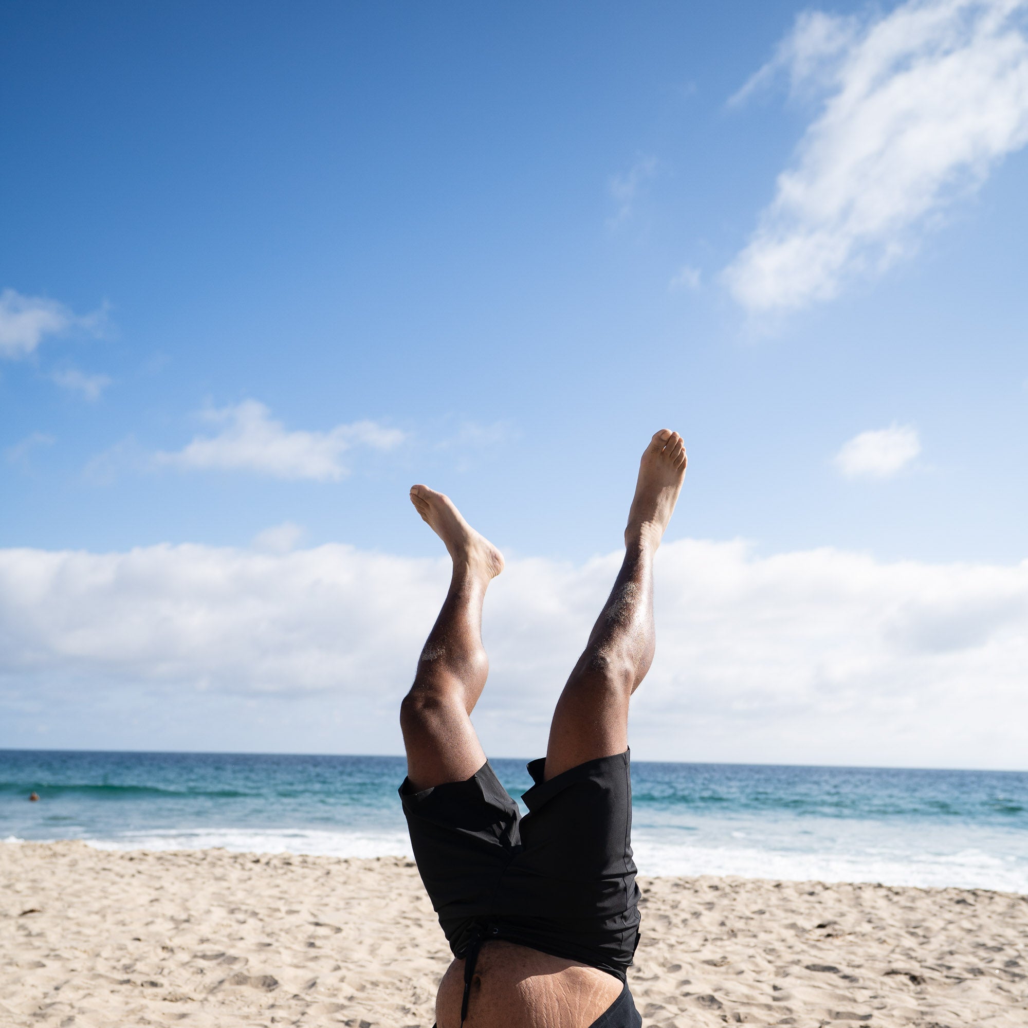 Person performing a handstand on a sandy beach, wearing Hoonigan Oil Slick board shorts against a clear blue sky.