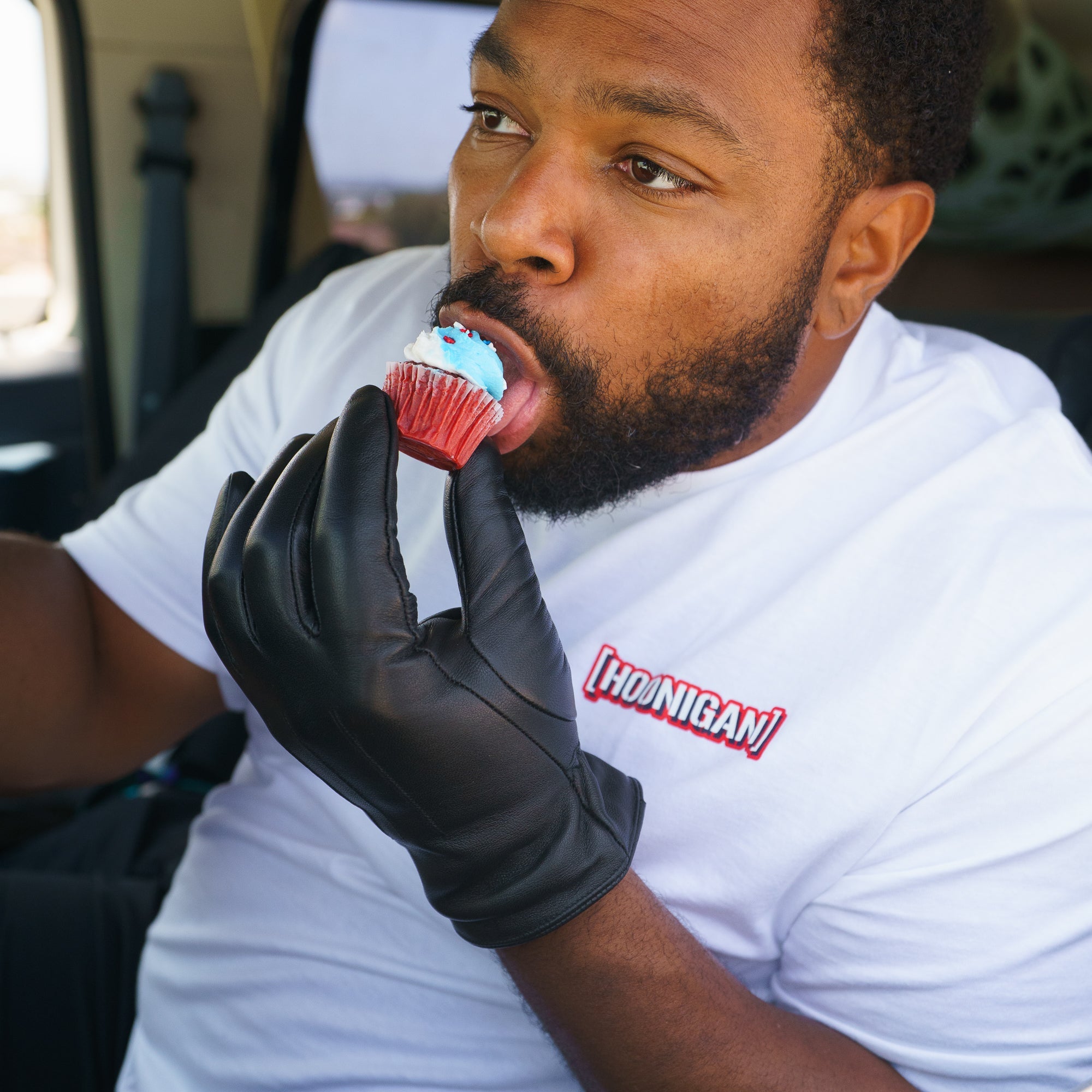 Man wearing a white Hoonigan T-shirt and black glove, licking a cupcake with blue frosting inside a vehicle.