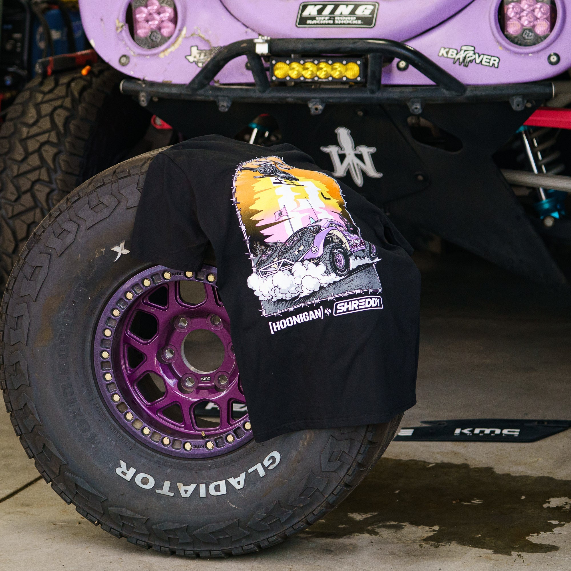 Hoonigan x Shreddy Desert Shred short sleeve t-shirt draped over a purple off-road tire in a garage setting.