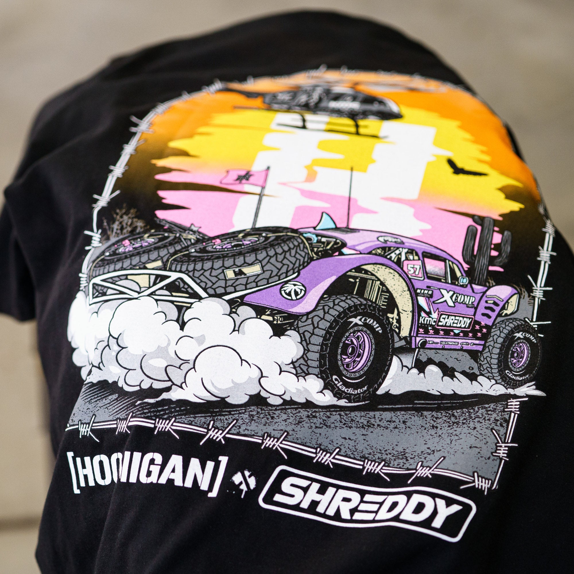 Black short sleeve t-shirt featuring vibrant graphic of a desert off-road vehicle with a sunset backdrop, showcasing Hoonigan x Shreddy collaboration.